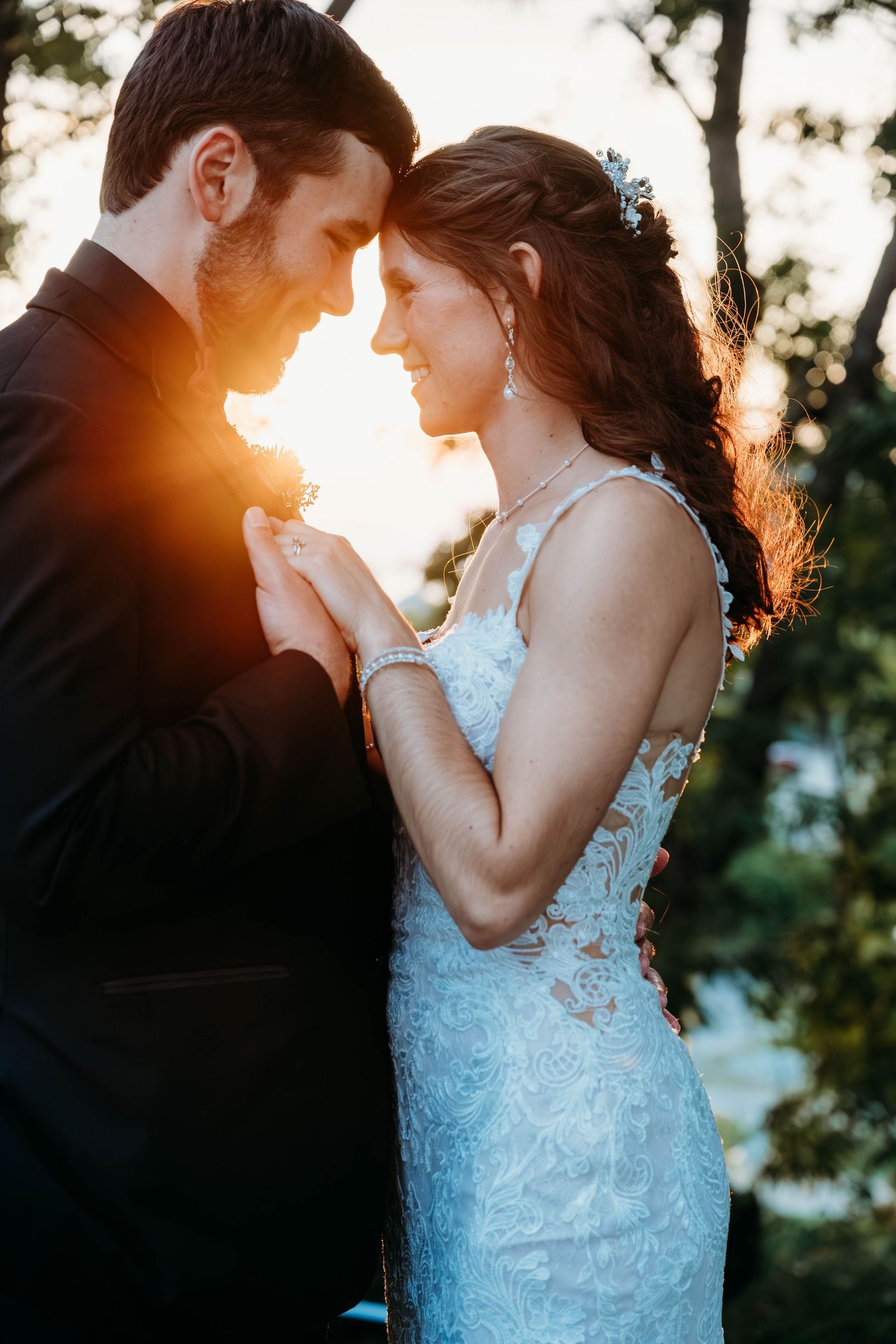 Bride and groom touch foreheads tenderly at golden hour sunset, holding hands in a romantic outdoor wedding portrait.