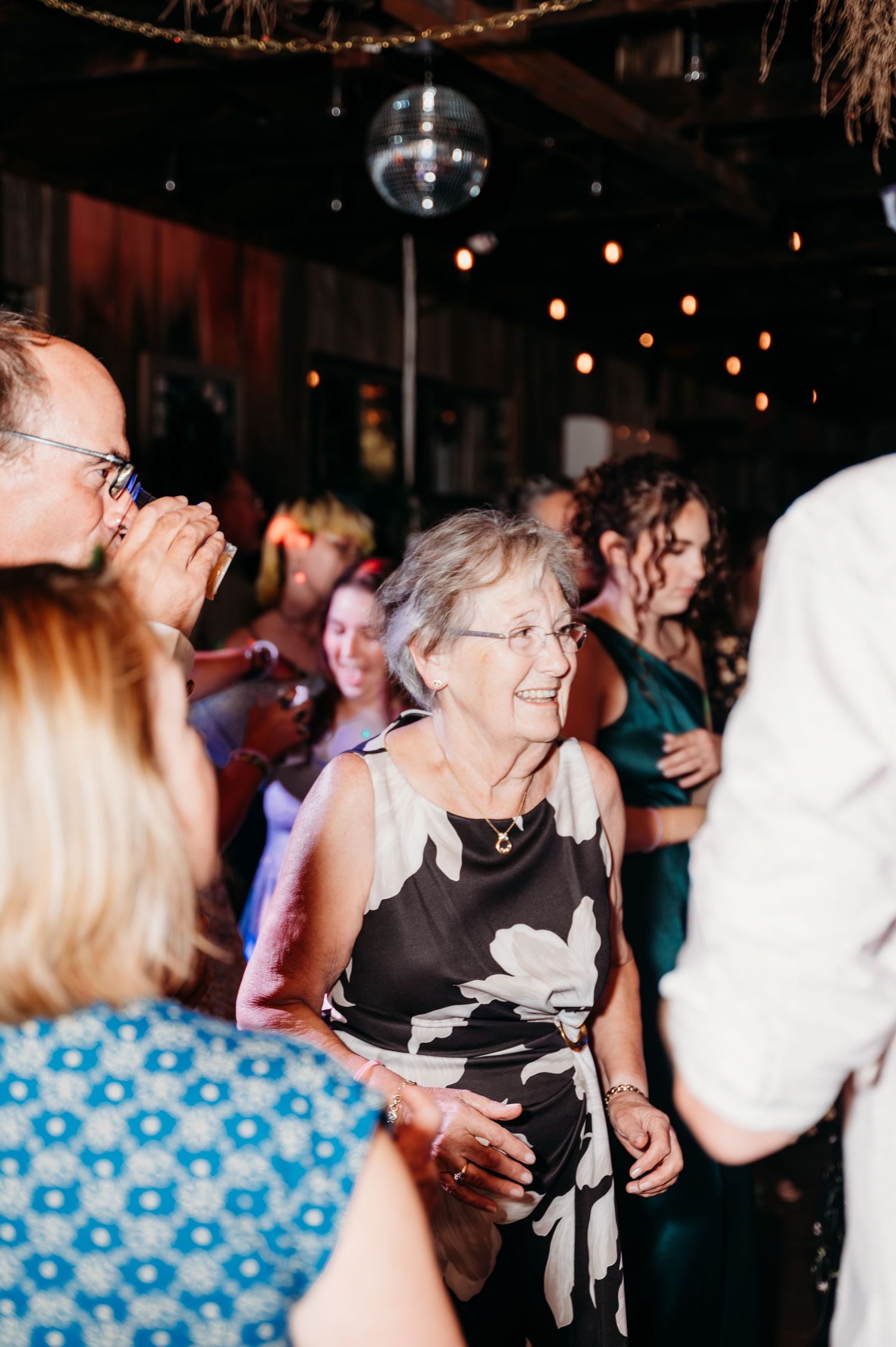 Elderly woman in floral dress dancing joyfully at a festive indoor party with string lights and disco ball.