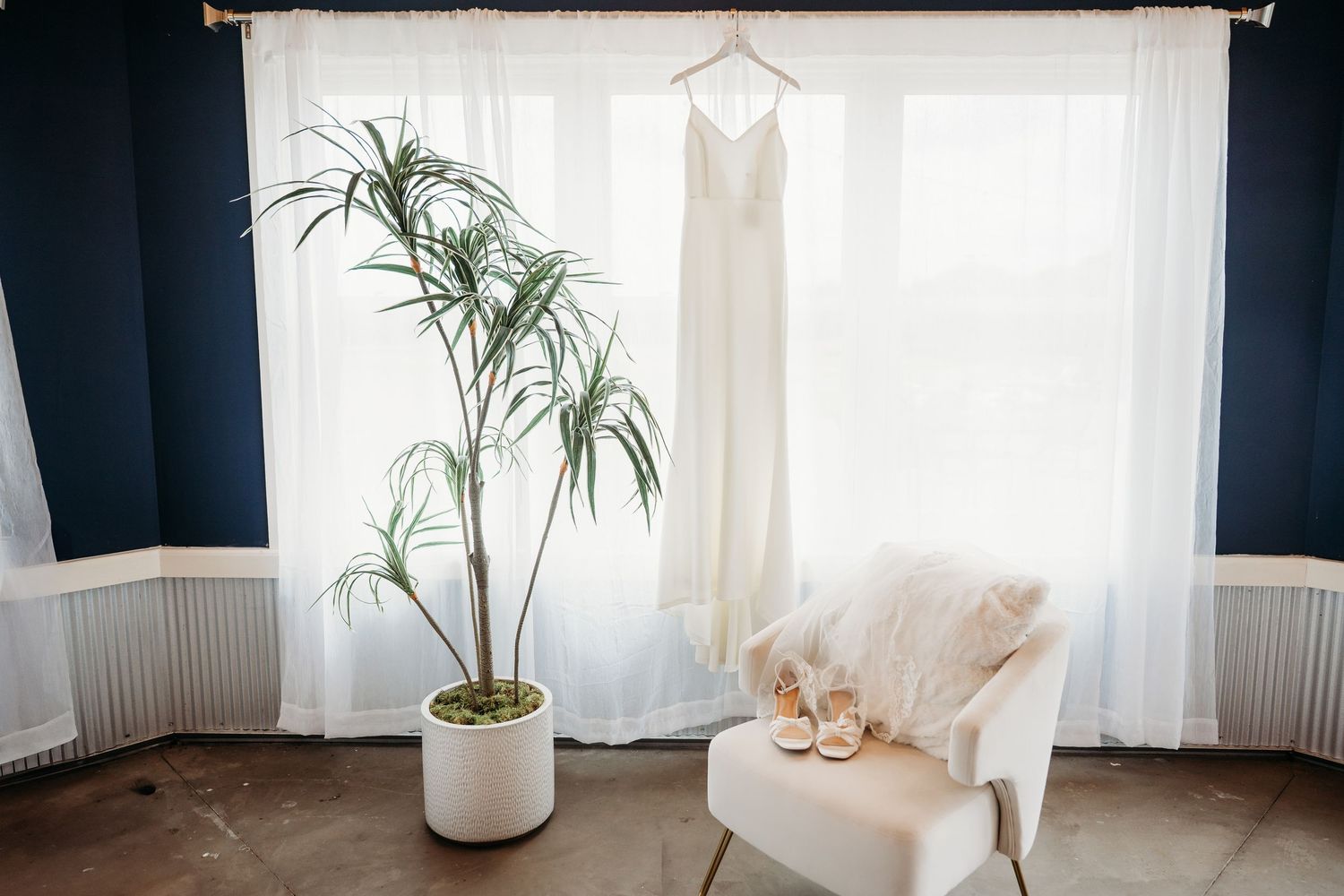 White wedding dress hanging by window with sheer curtains, tropical plant, and bridal heels on chair in elegant room.