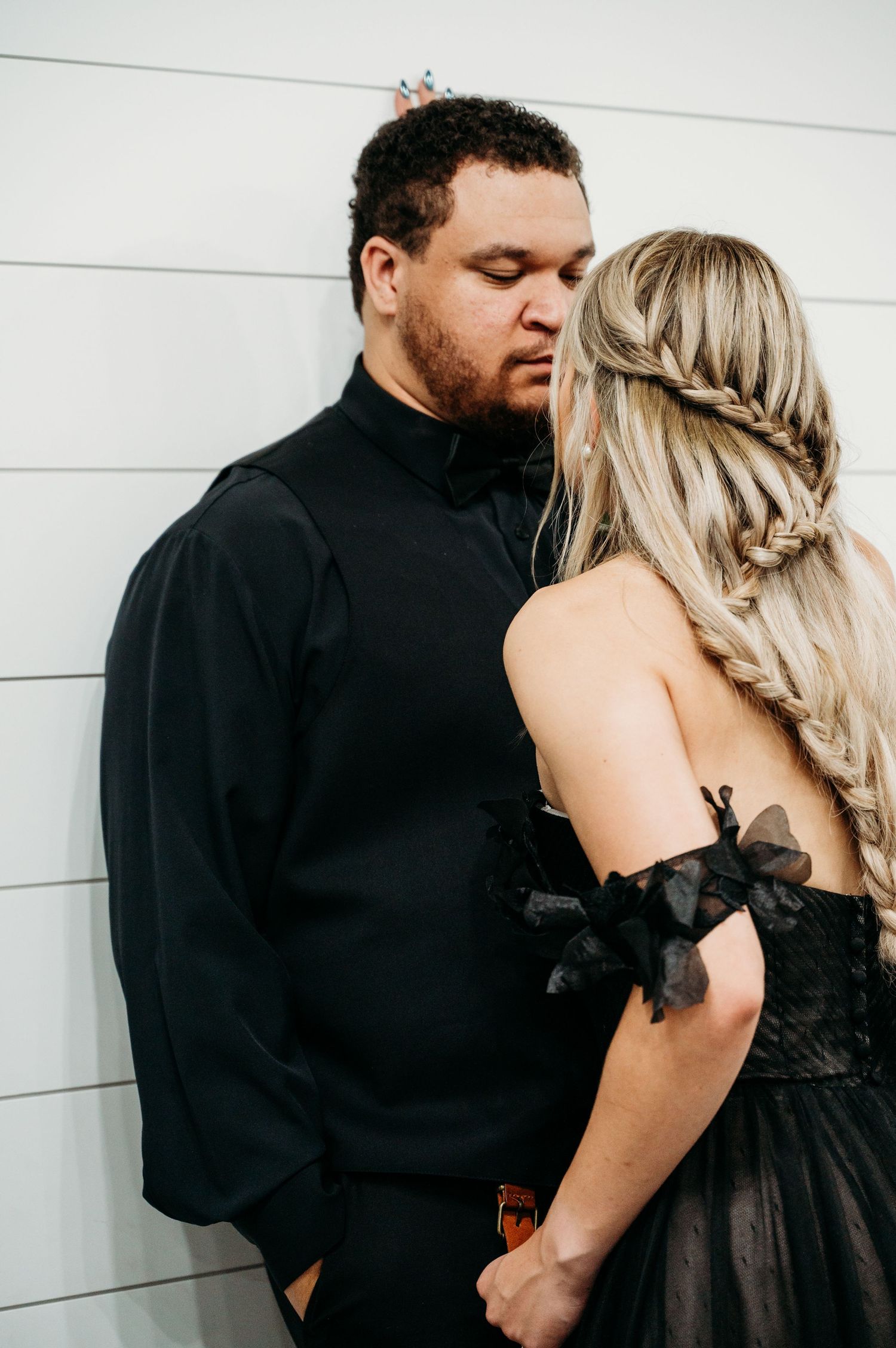 A romantic couple embraces against a white wall, with her in a black formal dress and him in a black turtleneck.