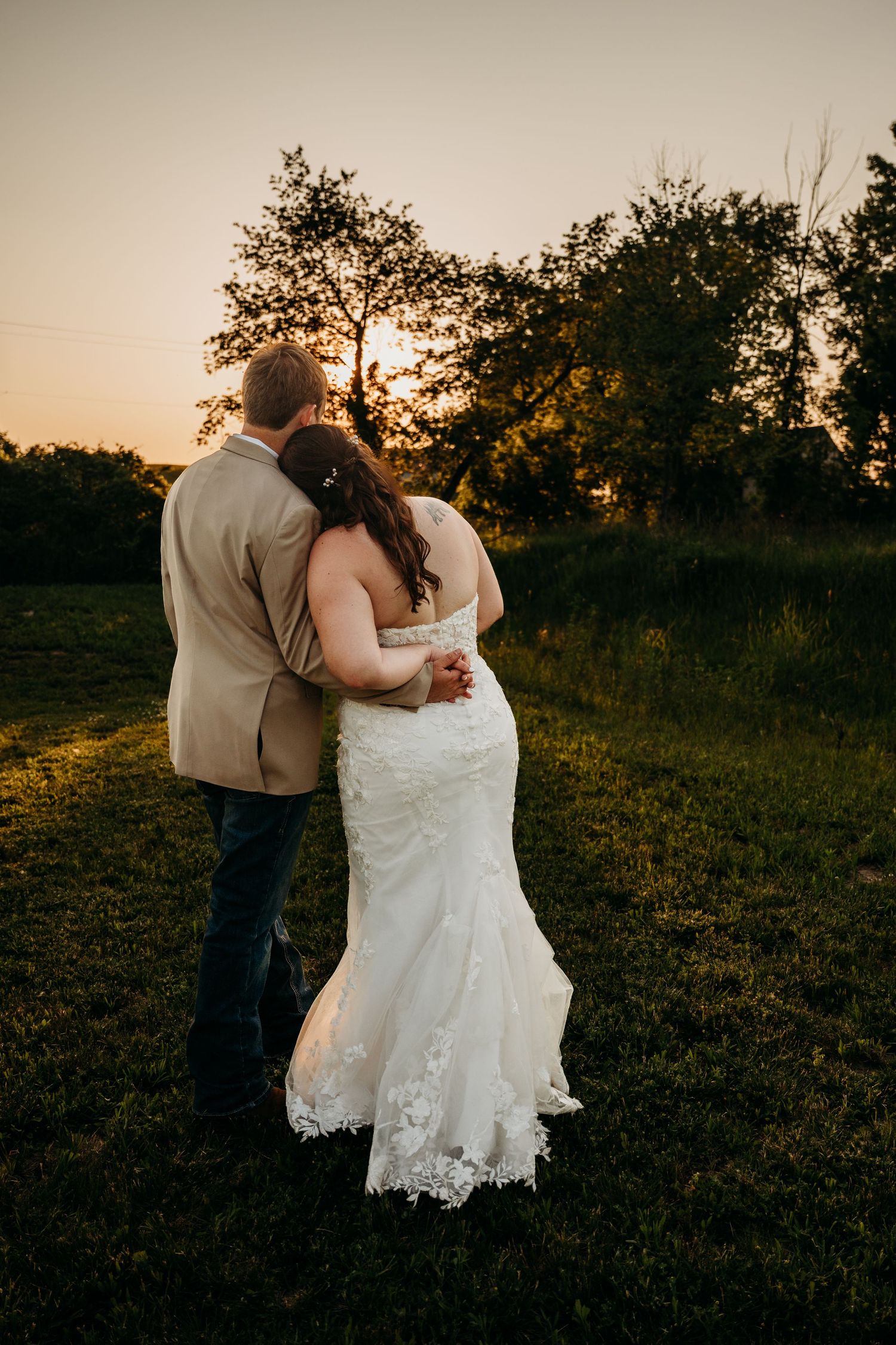 A romantic sunset wedding photo showing a couple embracing in a field with golden light filtering through trees.