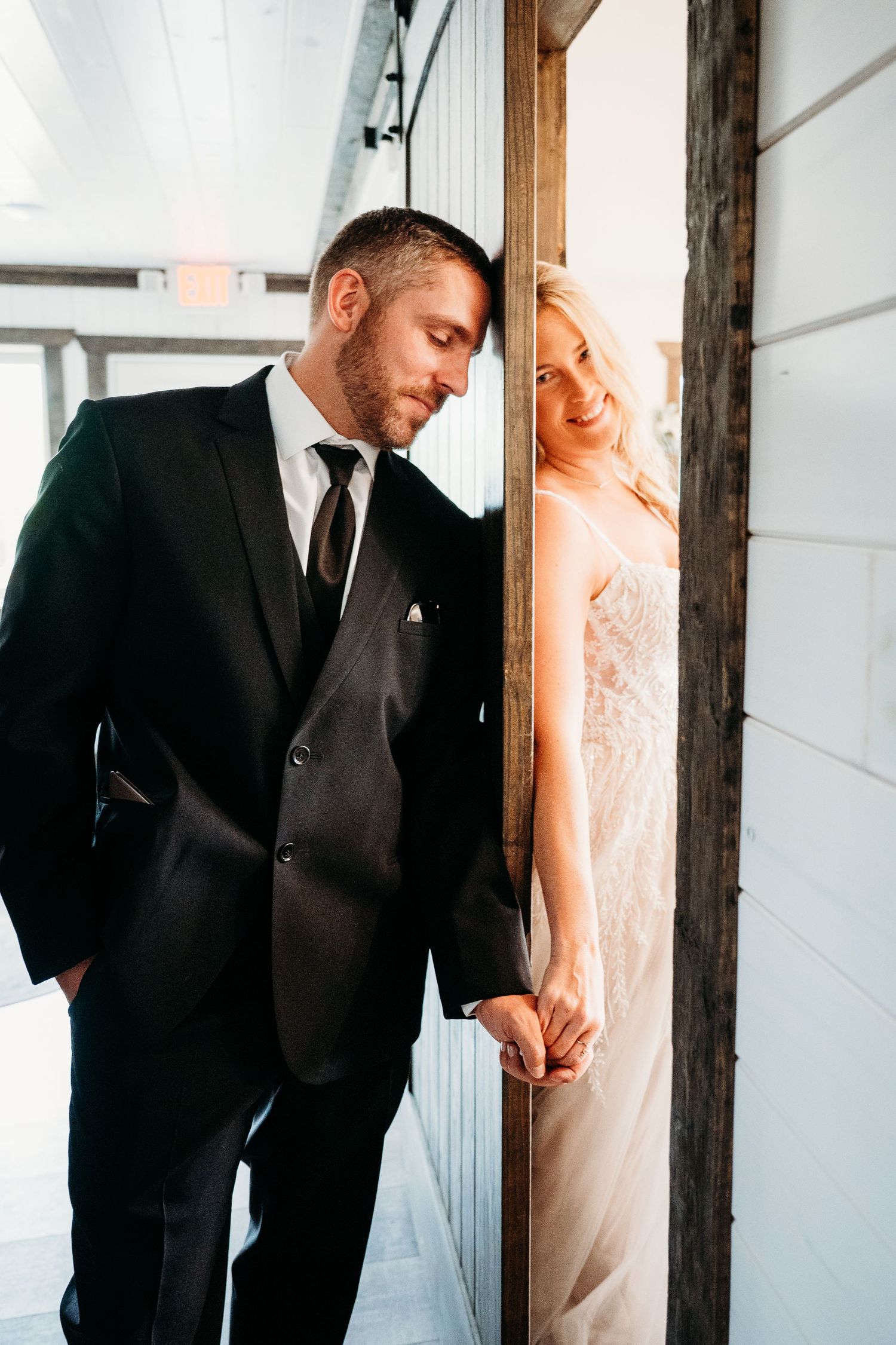A bride and groom share a tender moment while separated by a doorway, with the bride in white and groom in black suit.