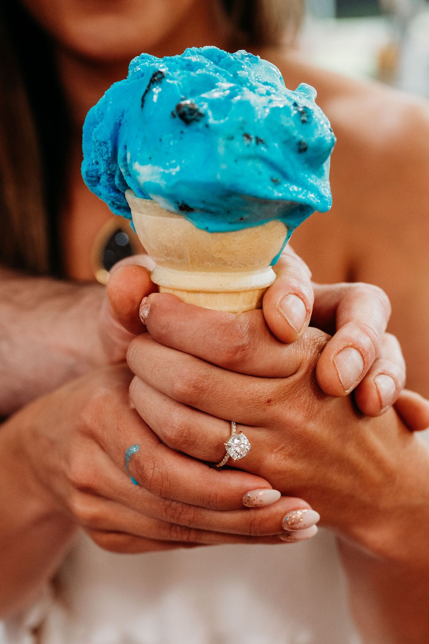 Hands holding a bright blue ice cream sandwich with an engagement ring visible.