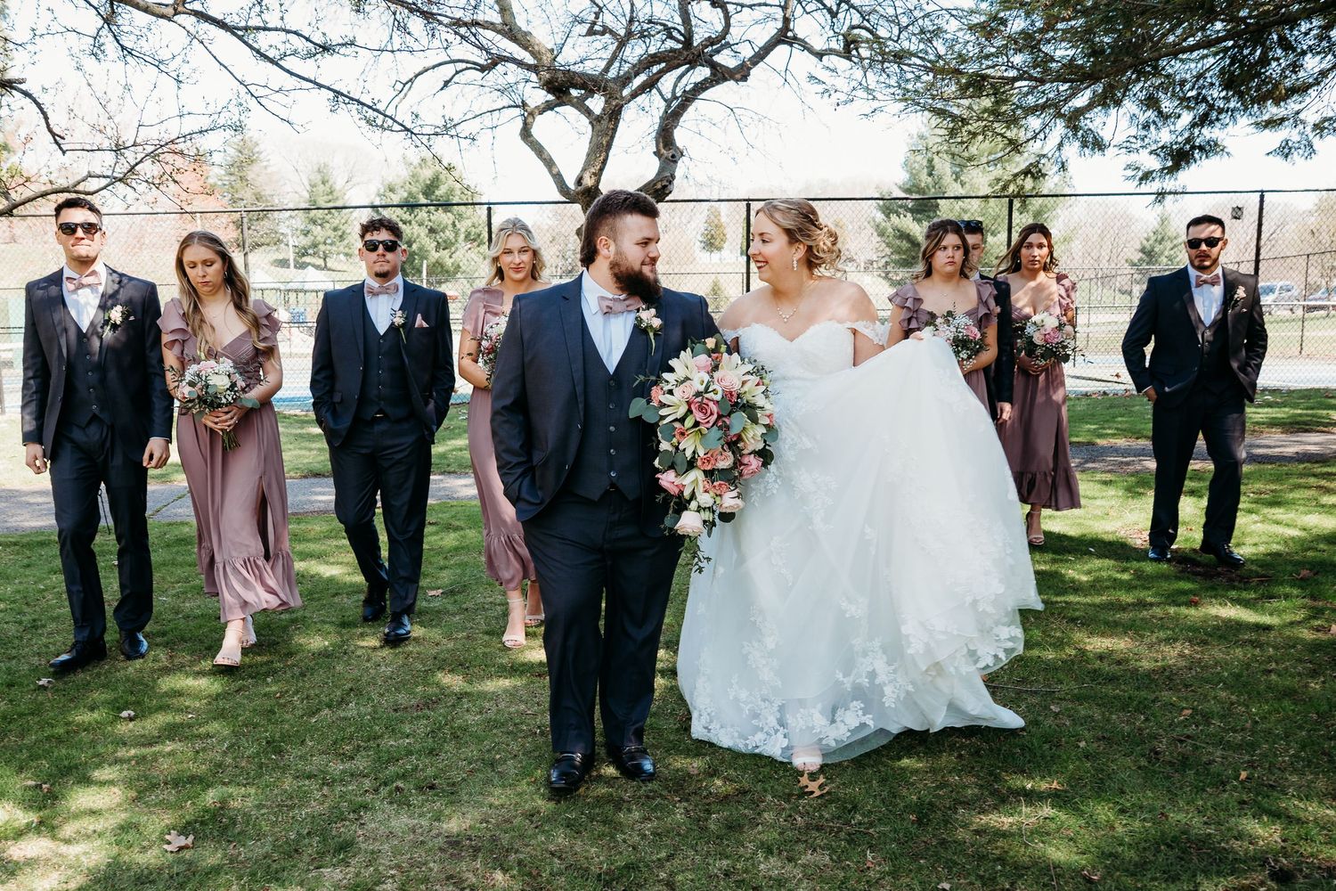 A wedding party in formal attire stands on a grassy lawn with bridesmaids in mauve dresses and groomsmen in dark suits.