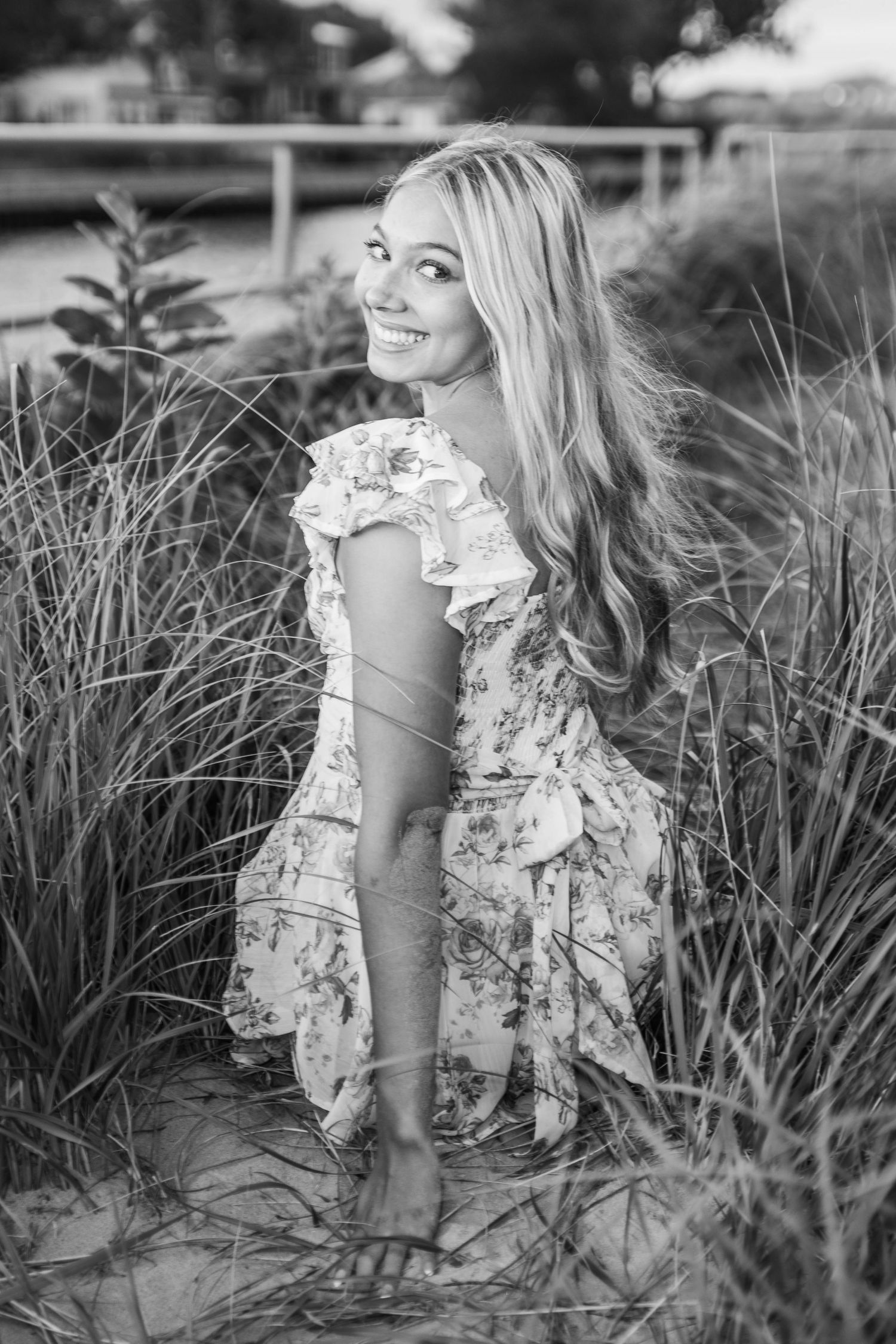 Black and white portrait in a floral dress posed among tall beach grasses.