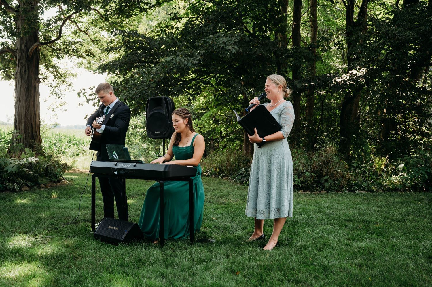 Outdoor wedding band performing with keyboard, guitar, and vocalist in a lush green garden setting.
