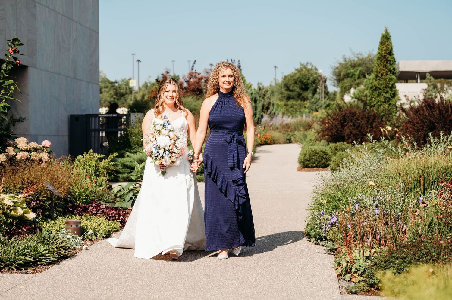 Bride in white gown walks with bridesmaid in navy dress through a garden path on a sunny wedding day.