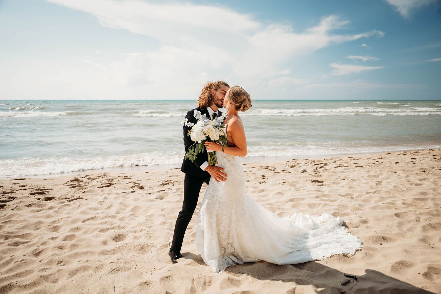 Bride and groom share a romantic kiss on a sandy beach with ocean waves and blue sky in the background.