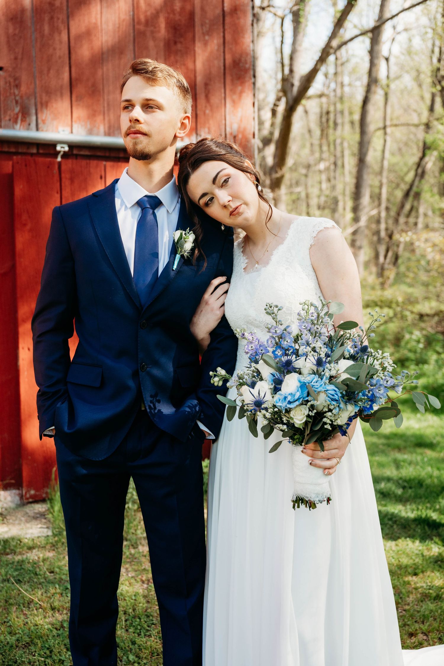 Bride in white gown holding blue floral bouquet leans on groom in navy suit near red barn outdoors.