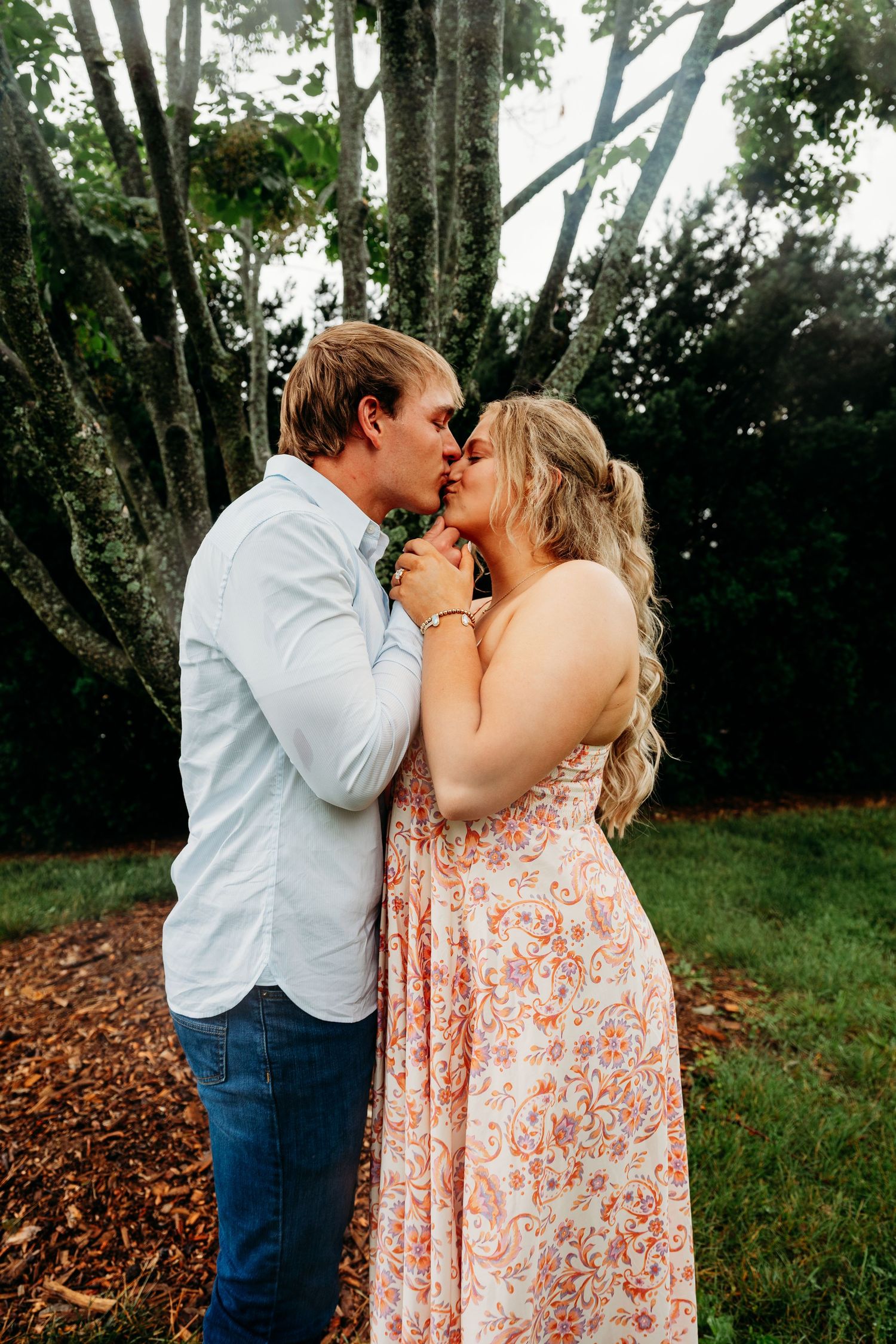 A couple shares a romantic kiss outdoors near large trees, woman in floral maxi dress, man in blue shirt and jeans.