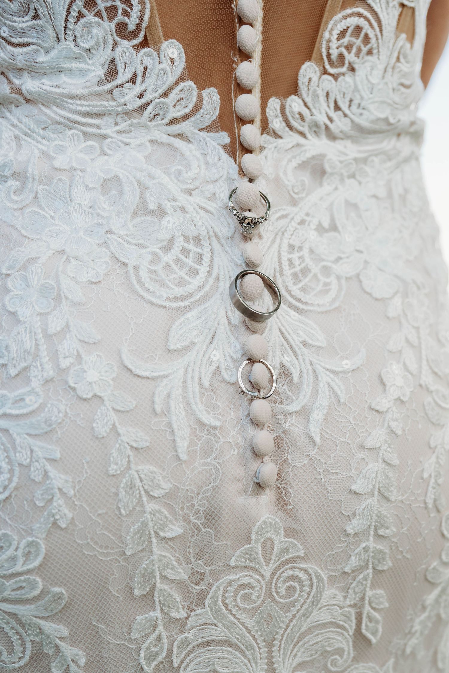 Wedding rings displayed on intricate white lace detail of a bridal gown with delicate button closures.