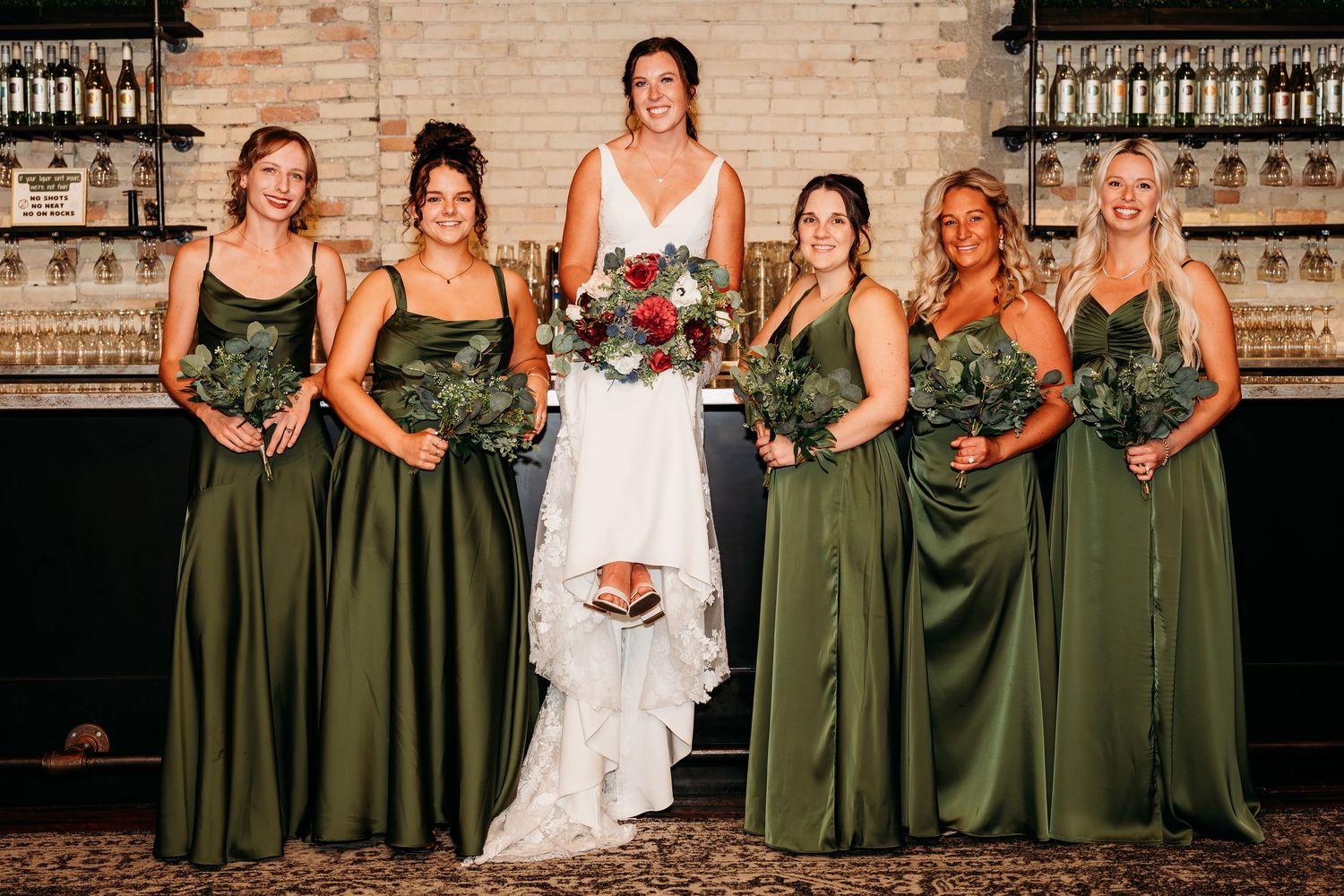 Wedding party poses in elegant olive green bridesmaid dresses and white bridal gown against rustic brick wall.