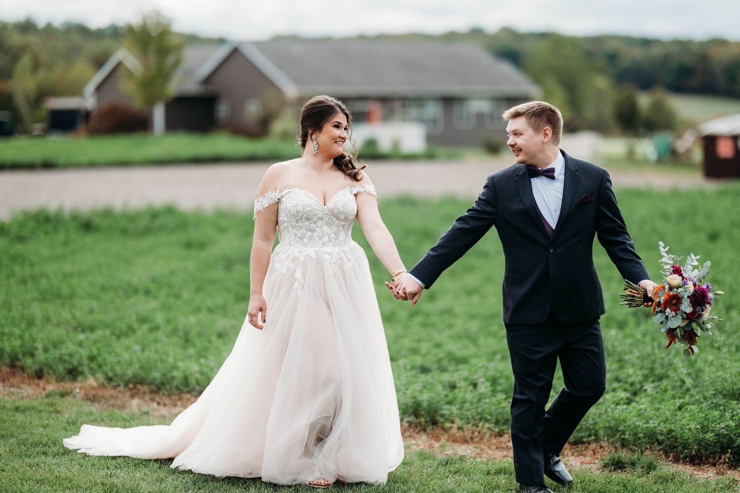 A couple walks hand in hand across a green field on their wedding day, with the bride in white and groom in black tuxedo.