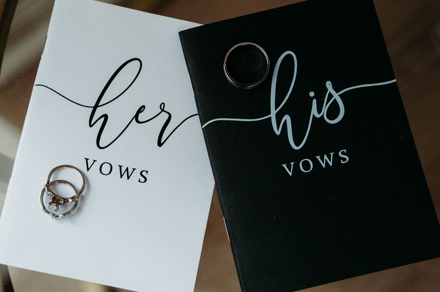 Wedding vow books in black and white with 'her' and 'his' calligraphy text alongside a wedding ring.