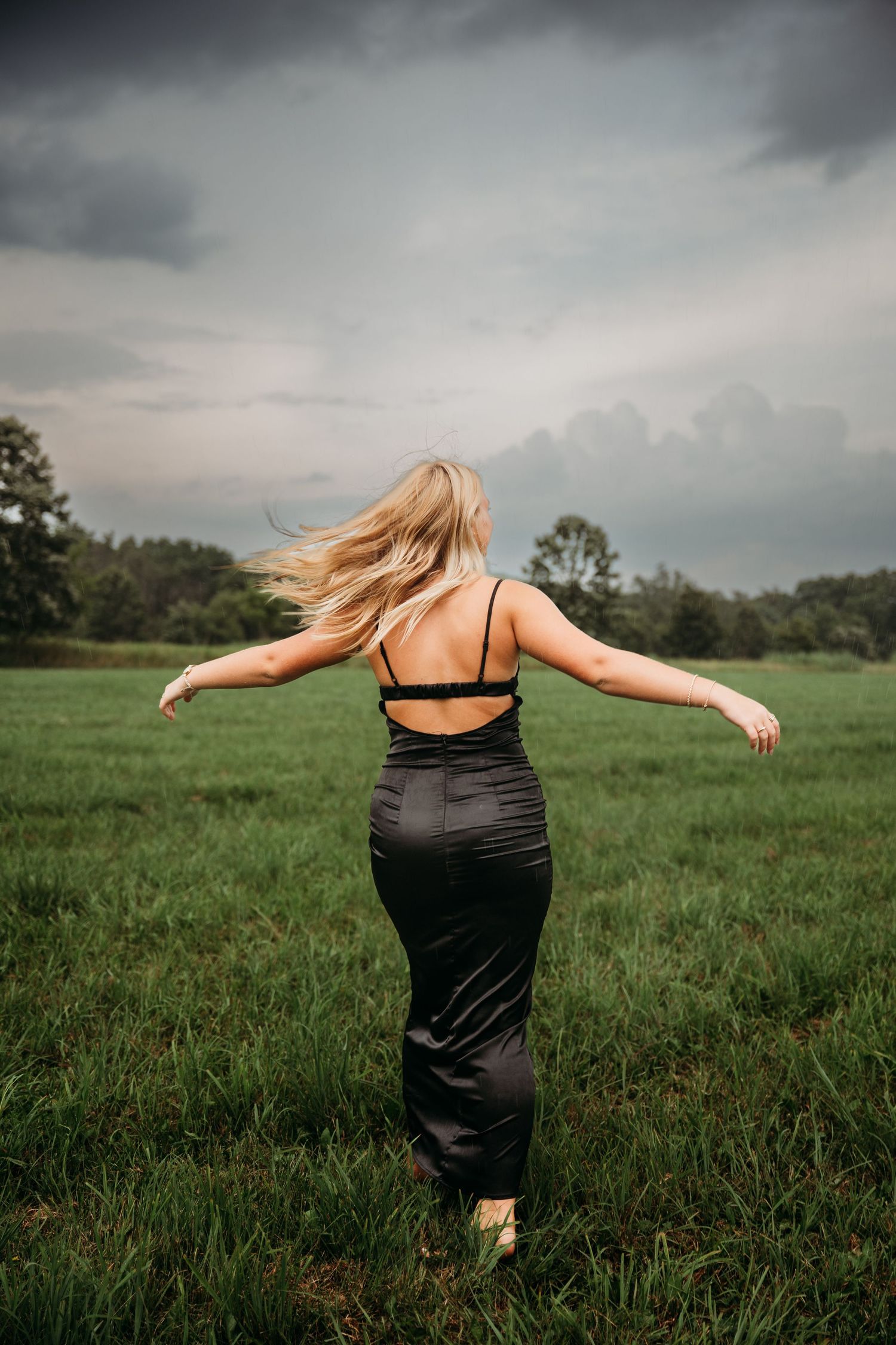 Figure in black dress spinning with arms outstretched in grassy field under cloudy sky.