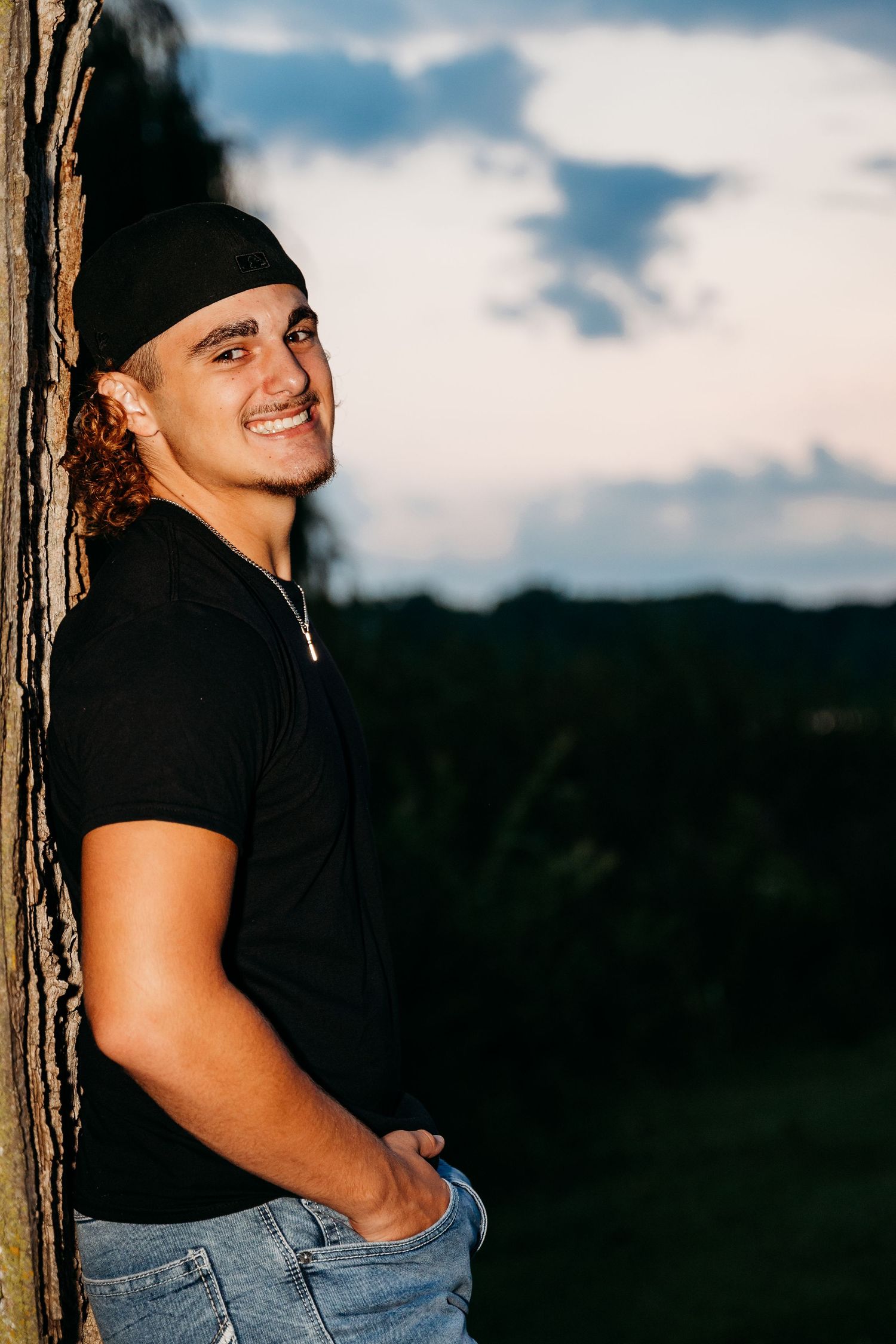 Person in black shirt and baseball cap leans against tree trunk with scenic view in background.