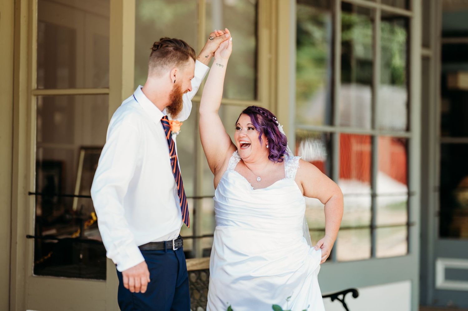 Wedding party members dance and celebrate during an outdoor reception with a glass-walled building in the background.