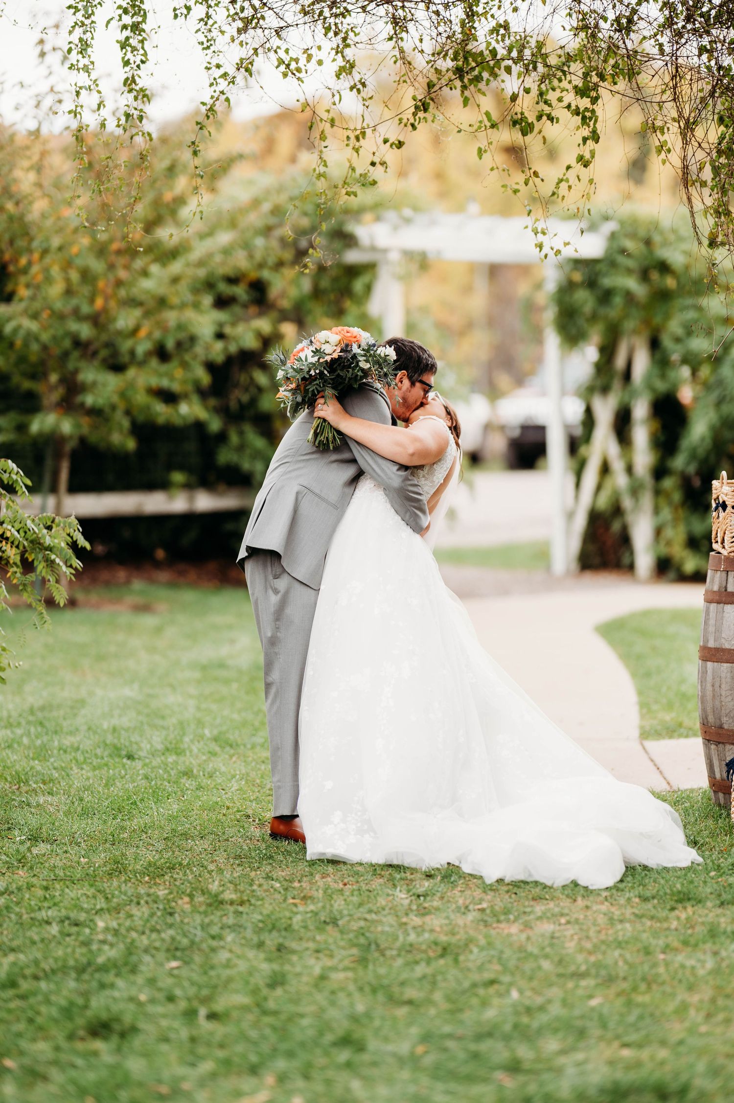 A romantic wedding couple embraces in a lush garden setting with autumn trees in the background.