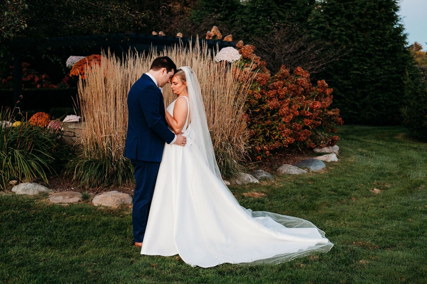 A romantic wedding couple embraces in a garden setting with tall ornamental grasses and autumn foliage.
