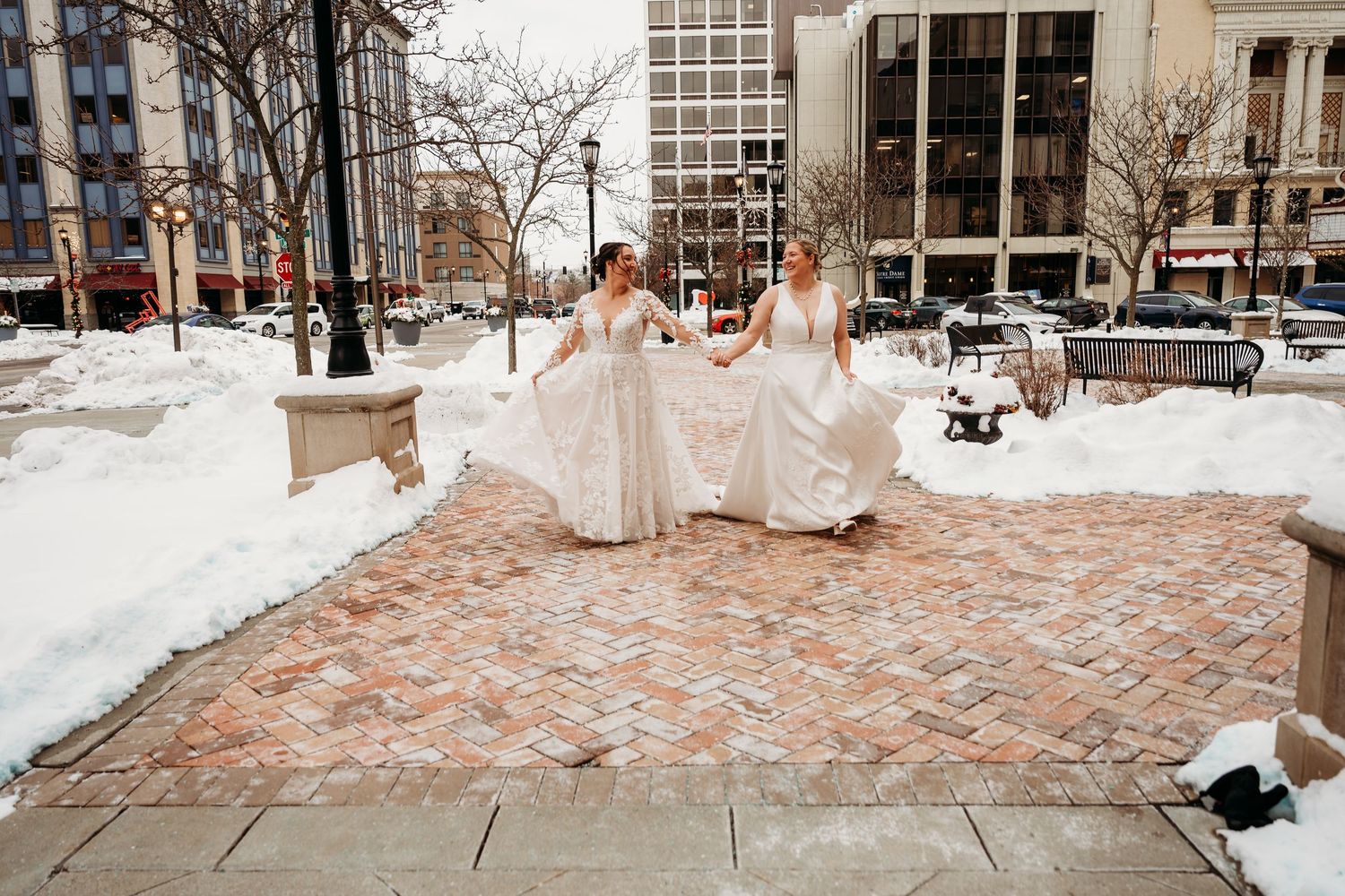 Two brides in white wedding dresses walk together on a snowy winter day along a brick pathway in an urban setting.