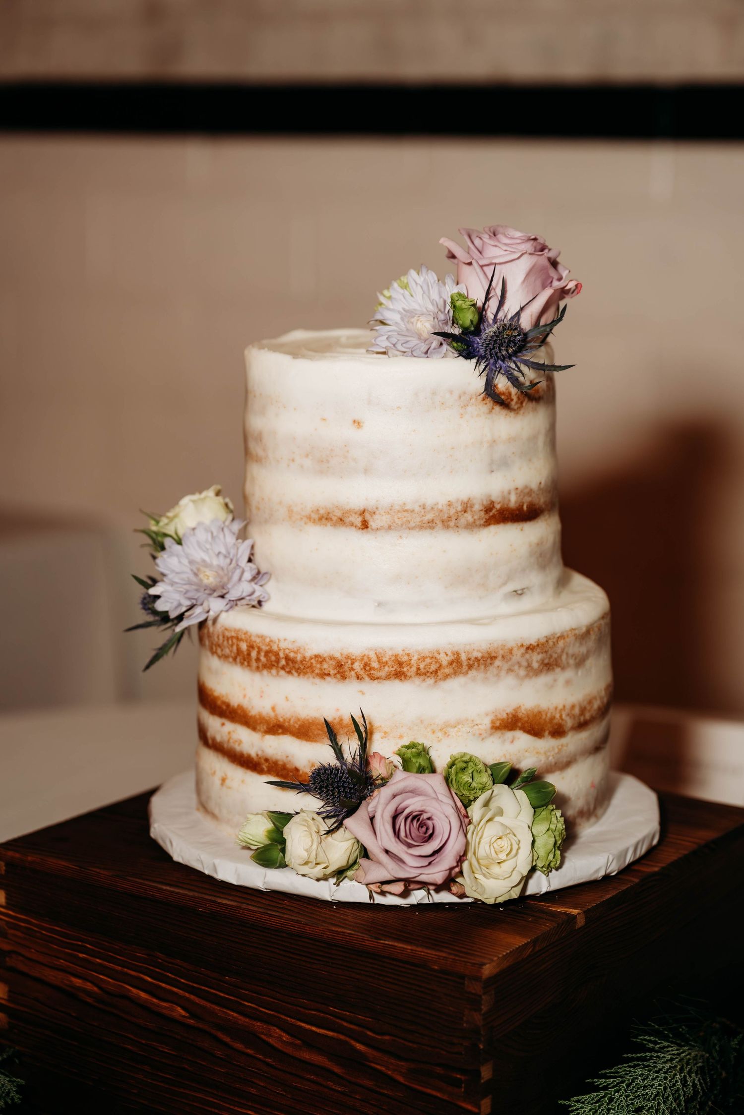 Rustic two-tier naked wedding cake decorated with pink roses and blue thistles on wooden display.