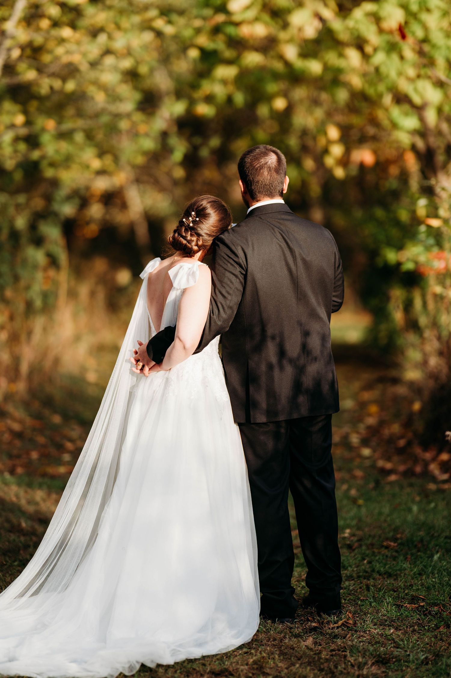 Bride in flowing white gown and groom in black suit embrace while walking away through an autumn garden setting.