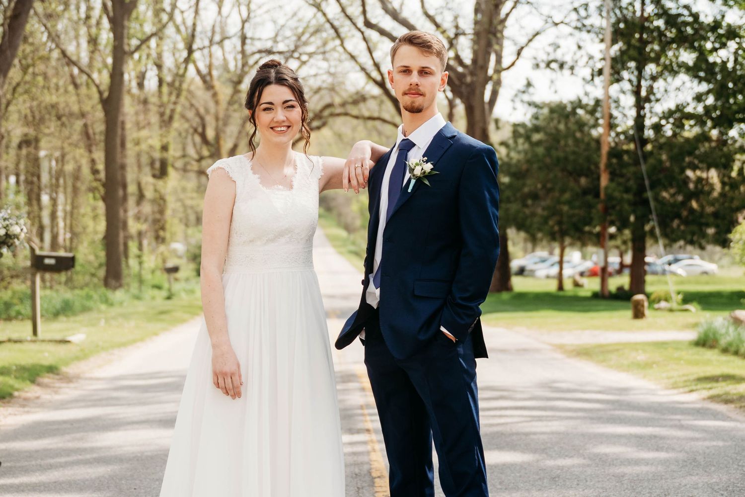 A bride in a white gown and groom in a navy suit pose together on a tree-lined path at their outdoor wedding.