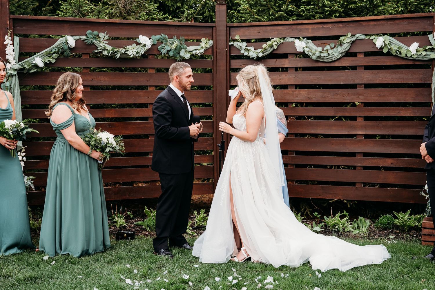 An outdoor wedding ceremony with a wooden fence backdrop adorned with white floral garlands and greenery.