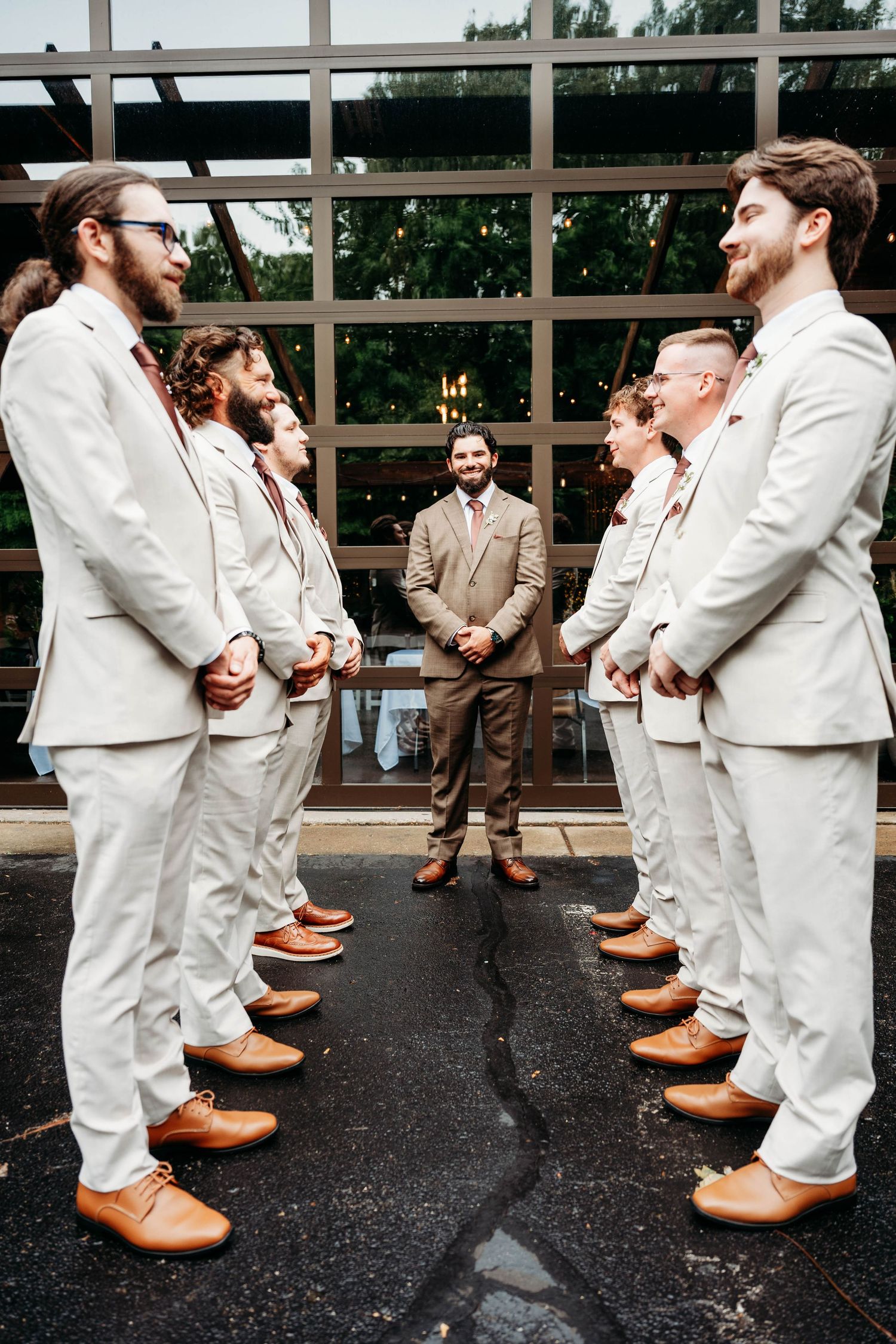 Groom stands facing groomsmen in cream suits and tan shoes outside a modern glass building.