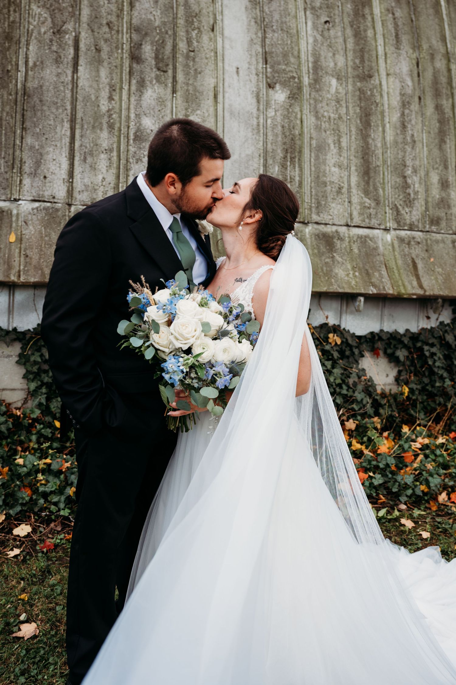 A romantic wedding couple shares a tender kiss against a rustic barn wall with climbing vines in the background.