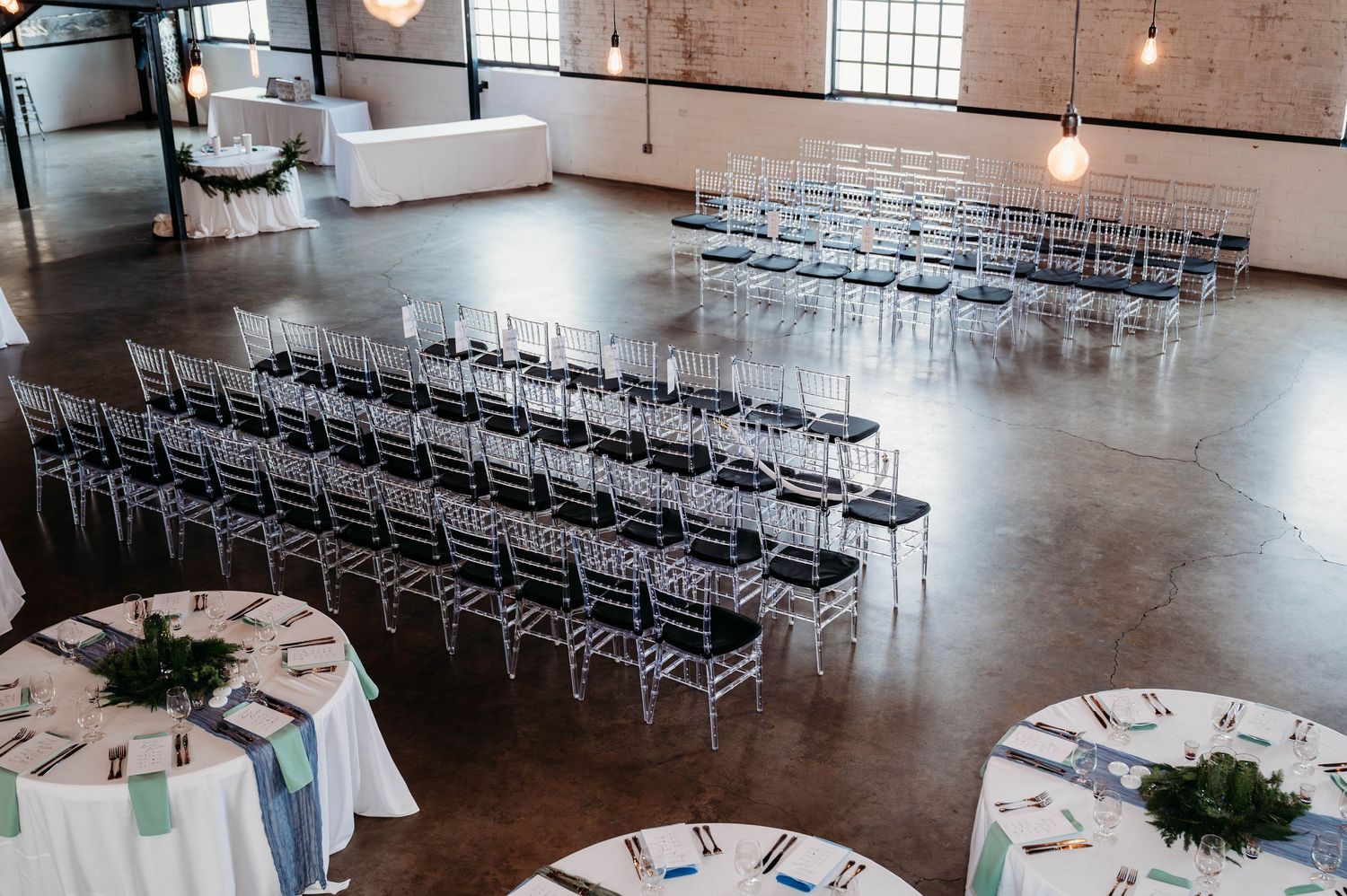 Modern industrial wedding venue with rows of silver chairs and round reception tables set up on polished concrete floors.