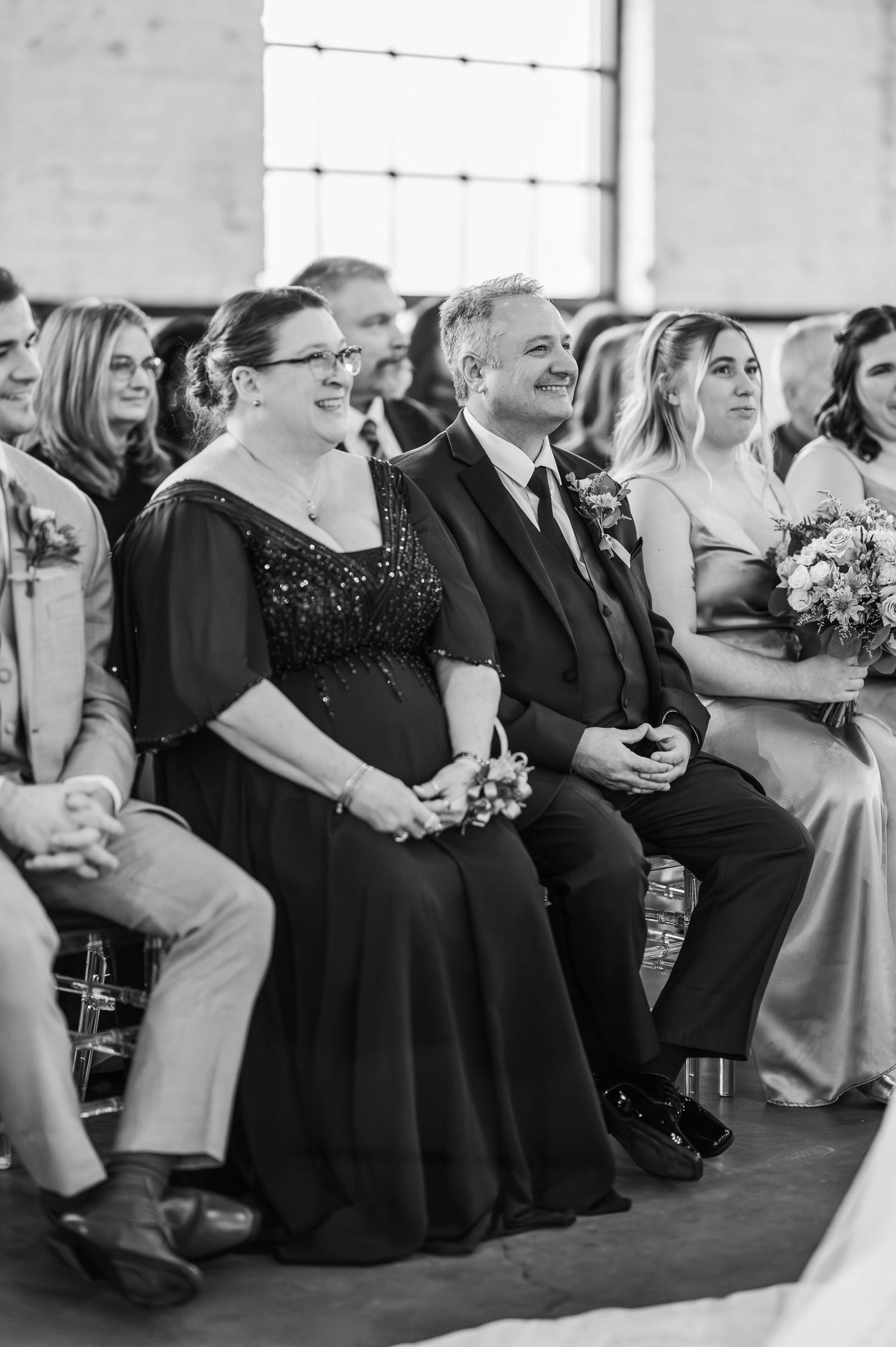 Wedding guests seated in formal attire watch an emotional ceremony at an indoor venue.