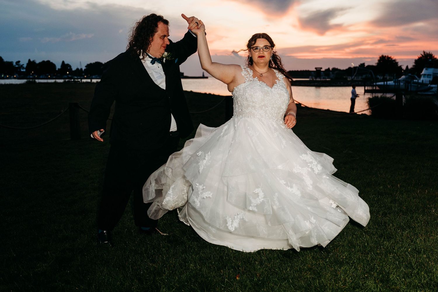 A couple in wedding attire dances in a field at sunset as their white dress twirls gracefully in the fading light.