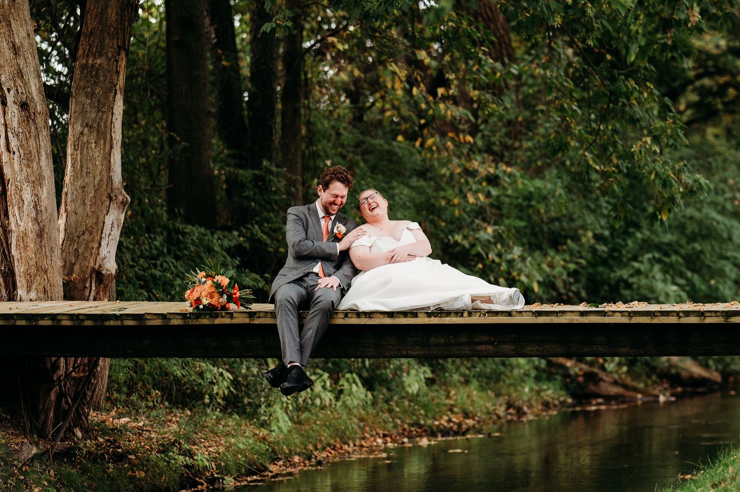 Newlyweds share a romantic moment on a rustic wooden bridge surrounded by autumn foliage in a park setting.