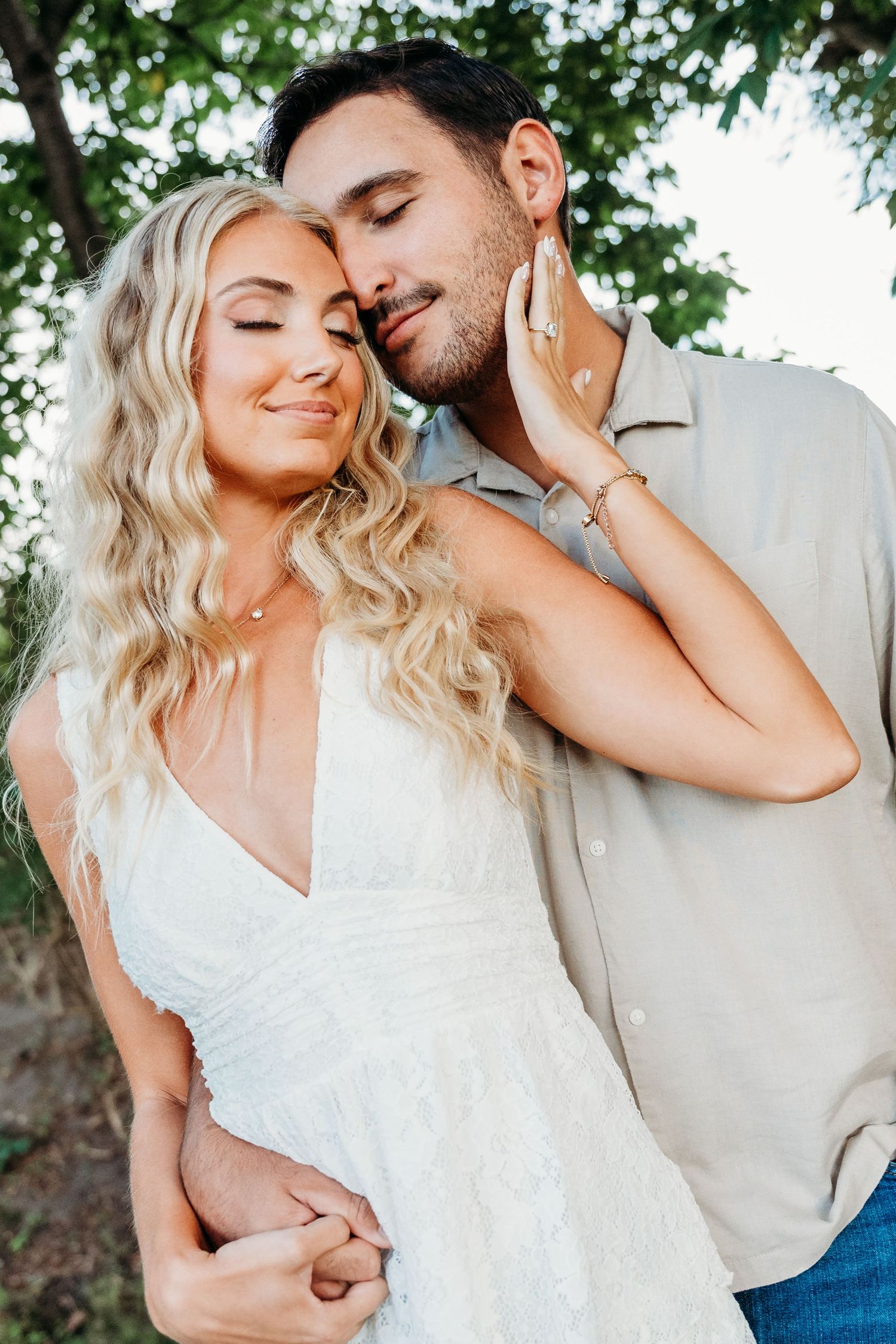 A romantic outdoor portrait shows an intimate moment between a couple, with her in white and him in beige against greenery.