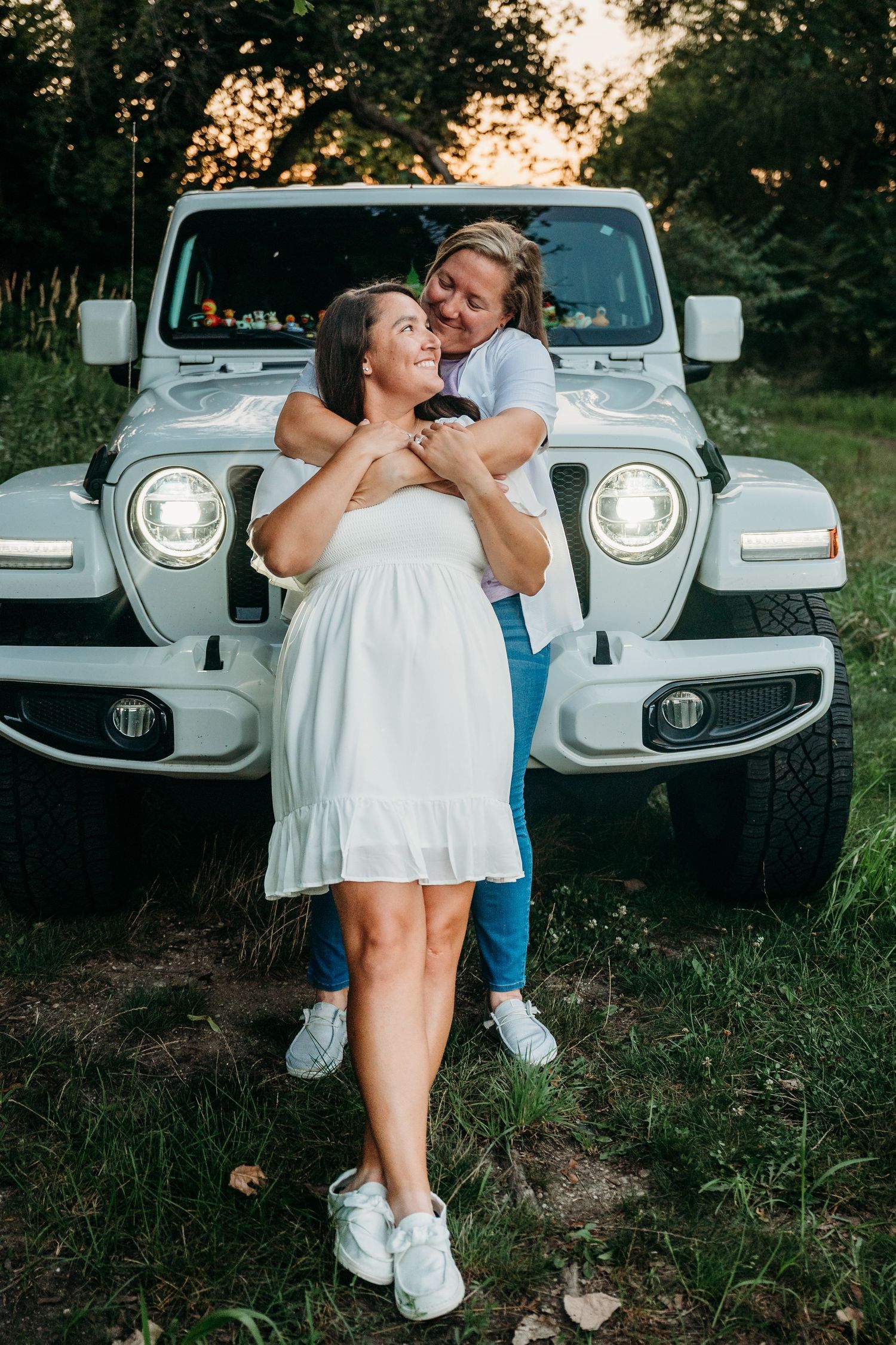 A couple shares a romantic moment in front of a silver Jeep at sunset in a wooded setting.