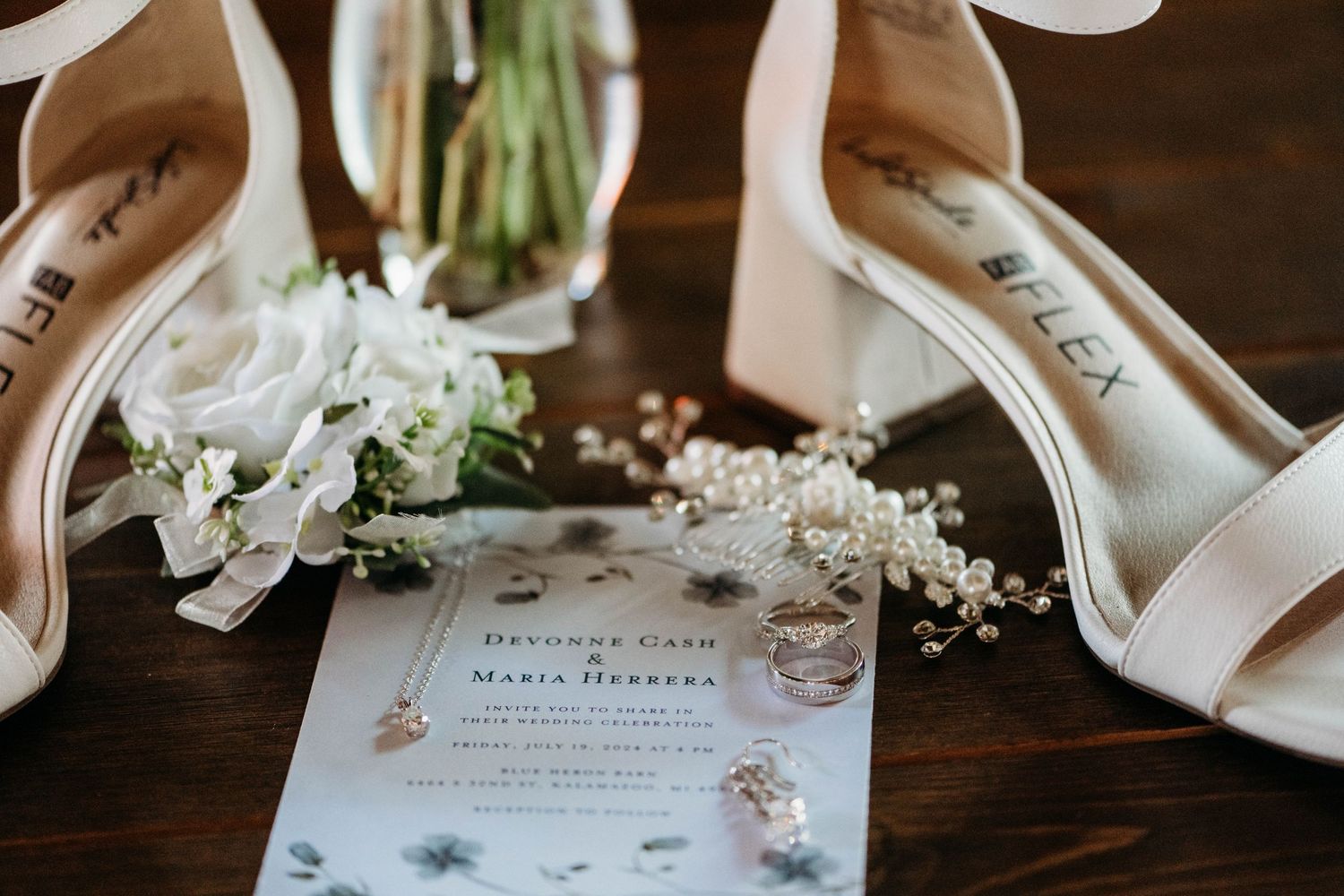 White wedding shoes and floral invitation card arranged on dark wooden surface with delicate white flowers.