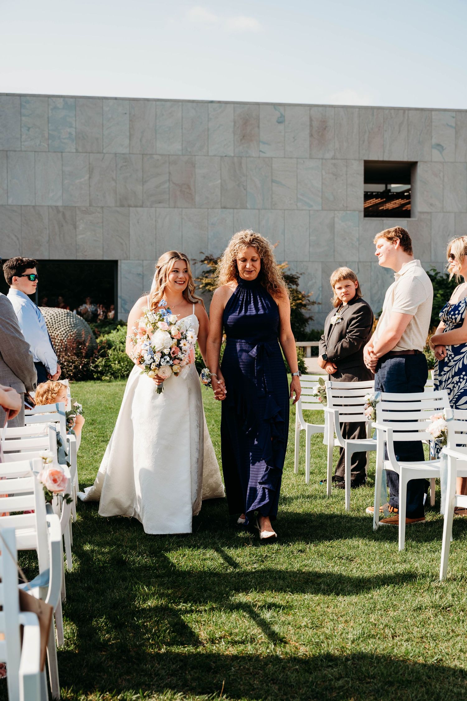 Bride in white gown walks down outdoor aisle with woman in navy dress, holding pastel bouquet at sunny wedding ceremony.