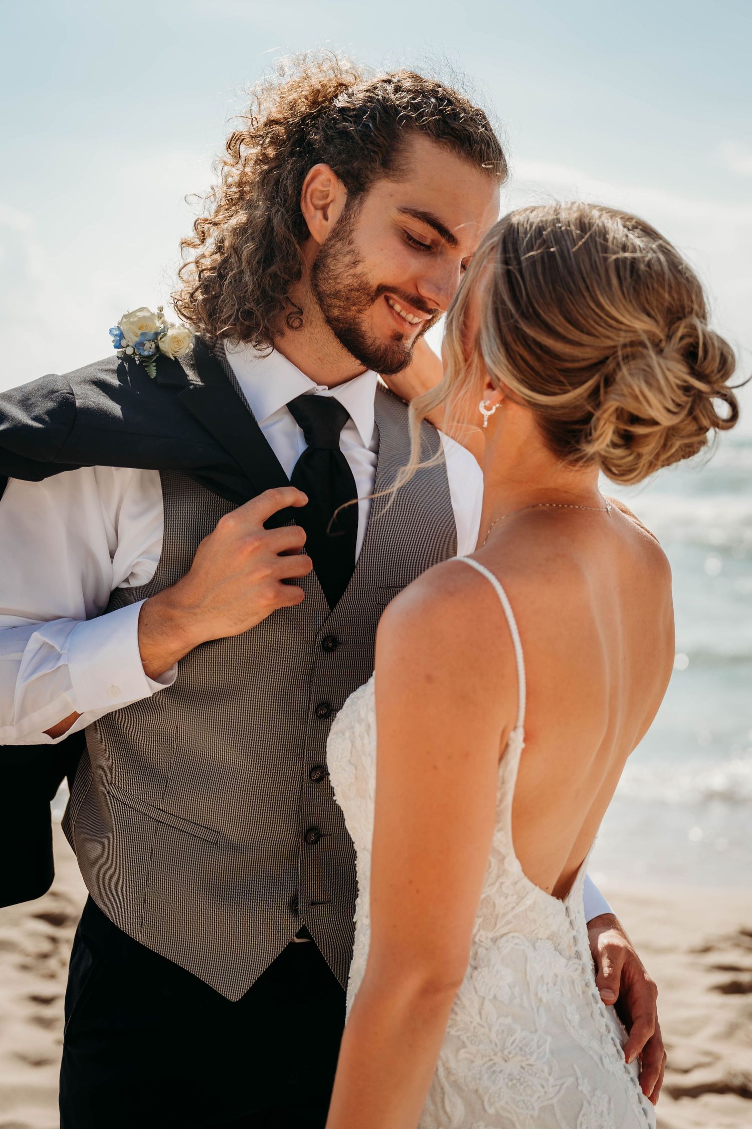 Smiling groom with curly hair leans in to kiss bride in lace dress on a sunny beach wedding day.