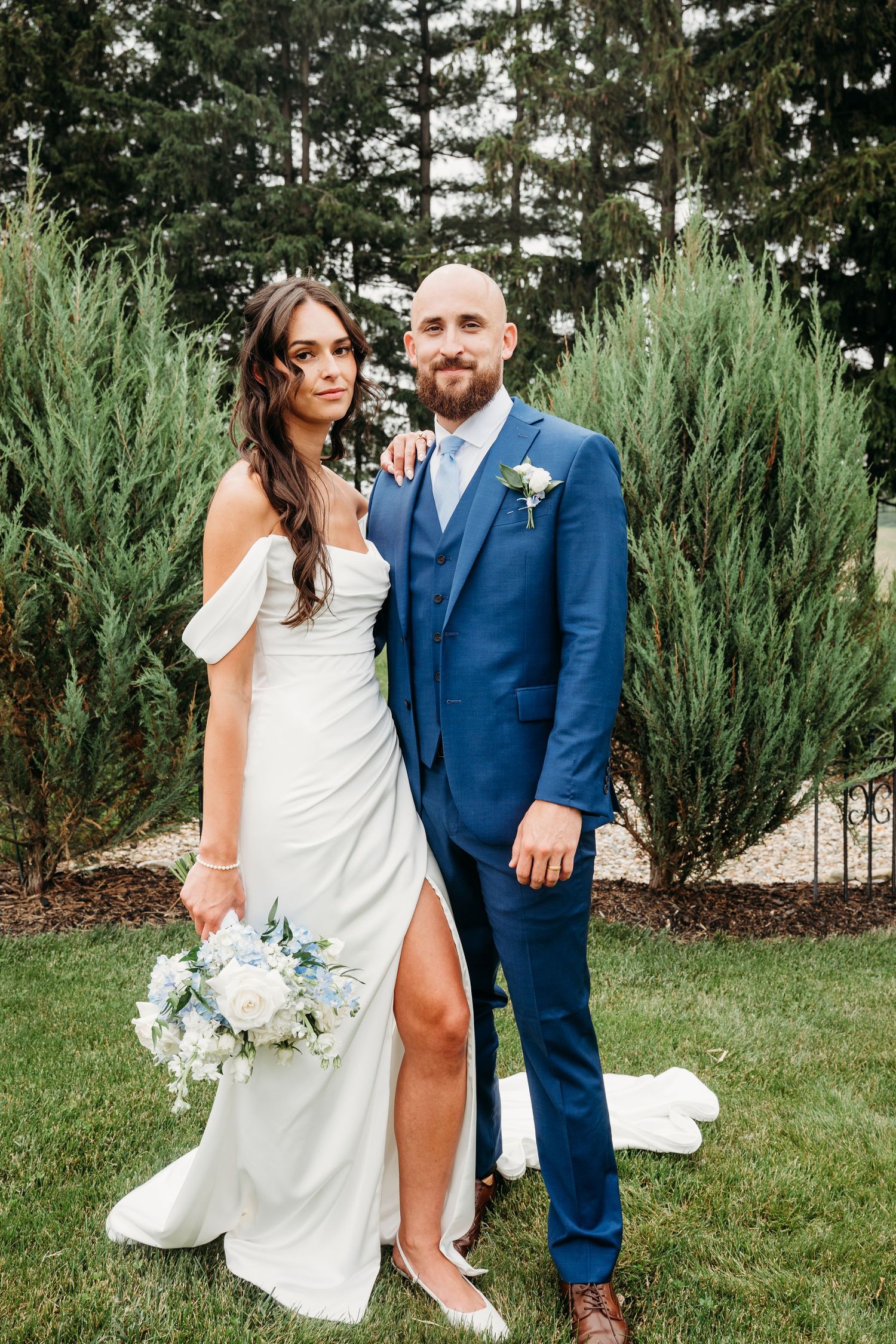 Bride in white gown holding bouquet poses with groom in blue suit outdoors on green lawn with evergreen trees. Res Les Farms