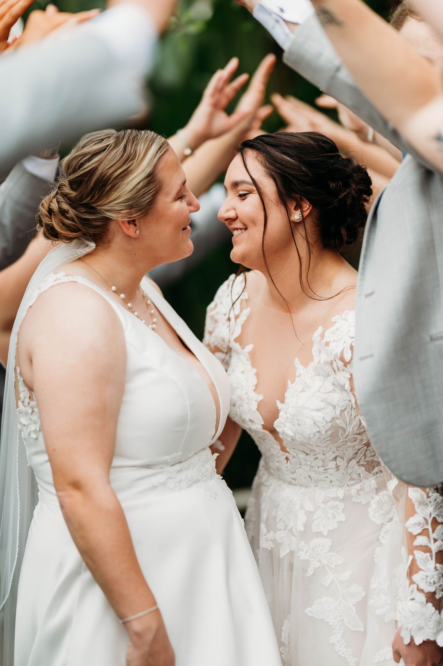 Two brides in white wedding gowns smile at each other during a joyful outdoor wedding celebration.