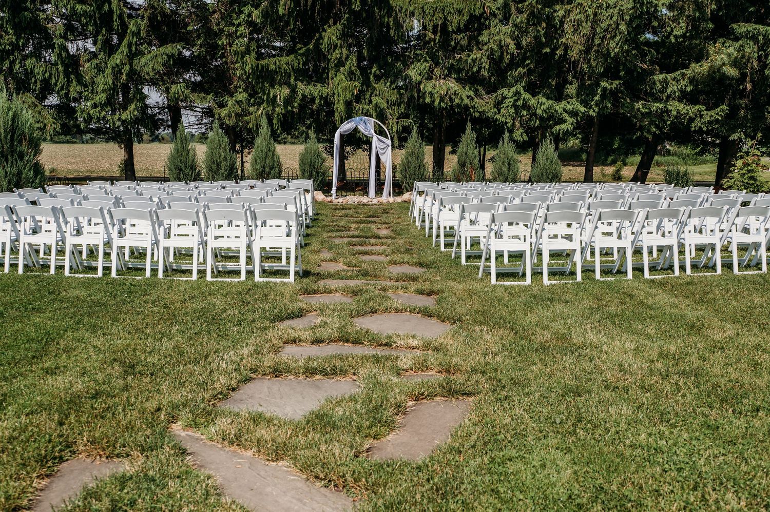 Outdoor wedding ceremony setup with white chairs lining a stone path leading to an arch surrounded by trees.