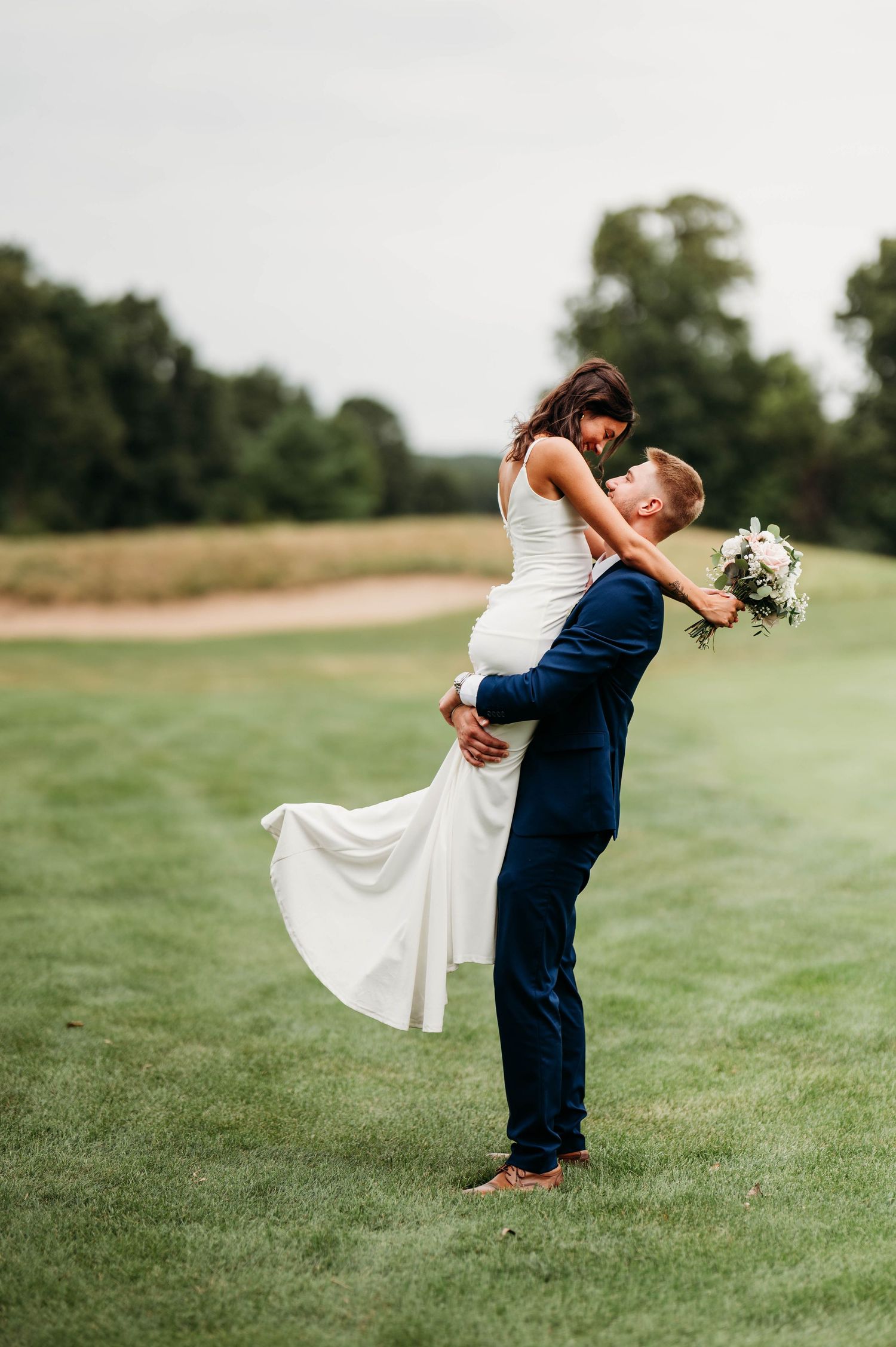 A romantic wedding couple shares a passionate kiss in a lush green field while the bride's dress flows in the breeze.