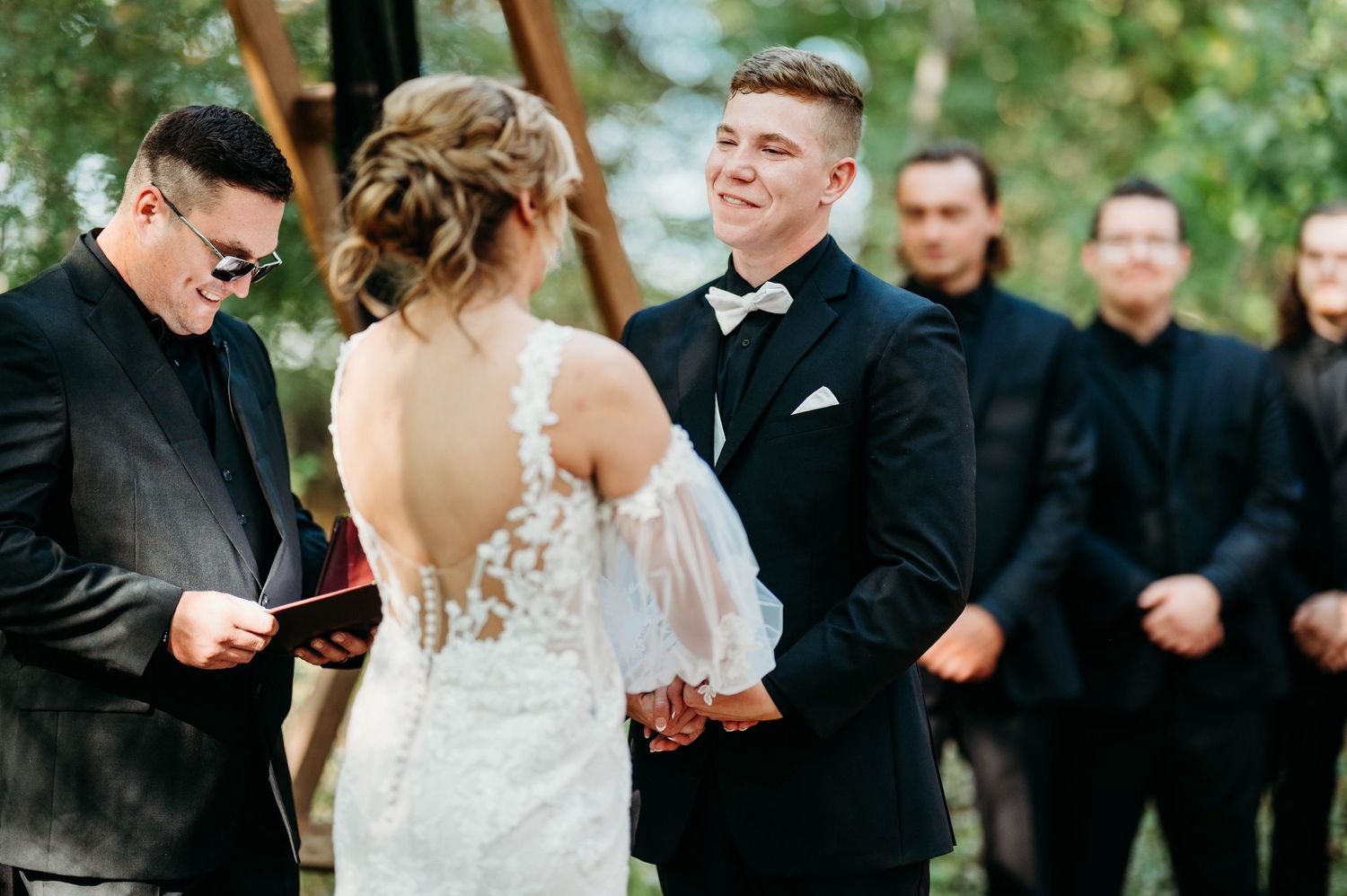 Wedding party members in black and white attire gather outdoors under trees during a celebration.