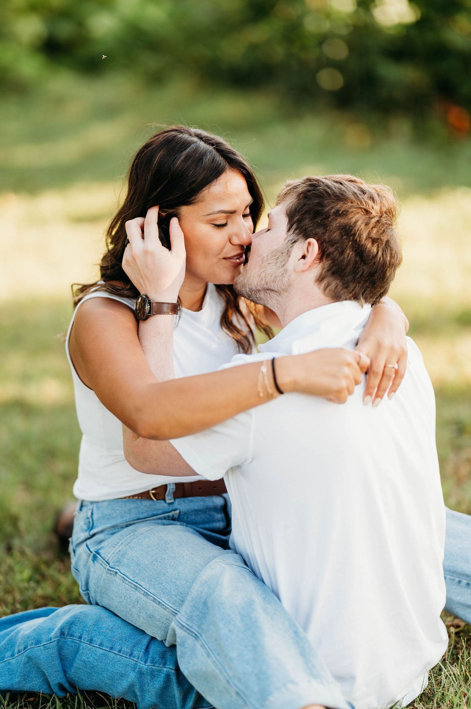 A couple shares an intimate moment while sitting together on grass in a sunlit outdoor setting.