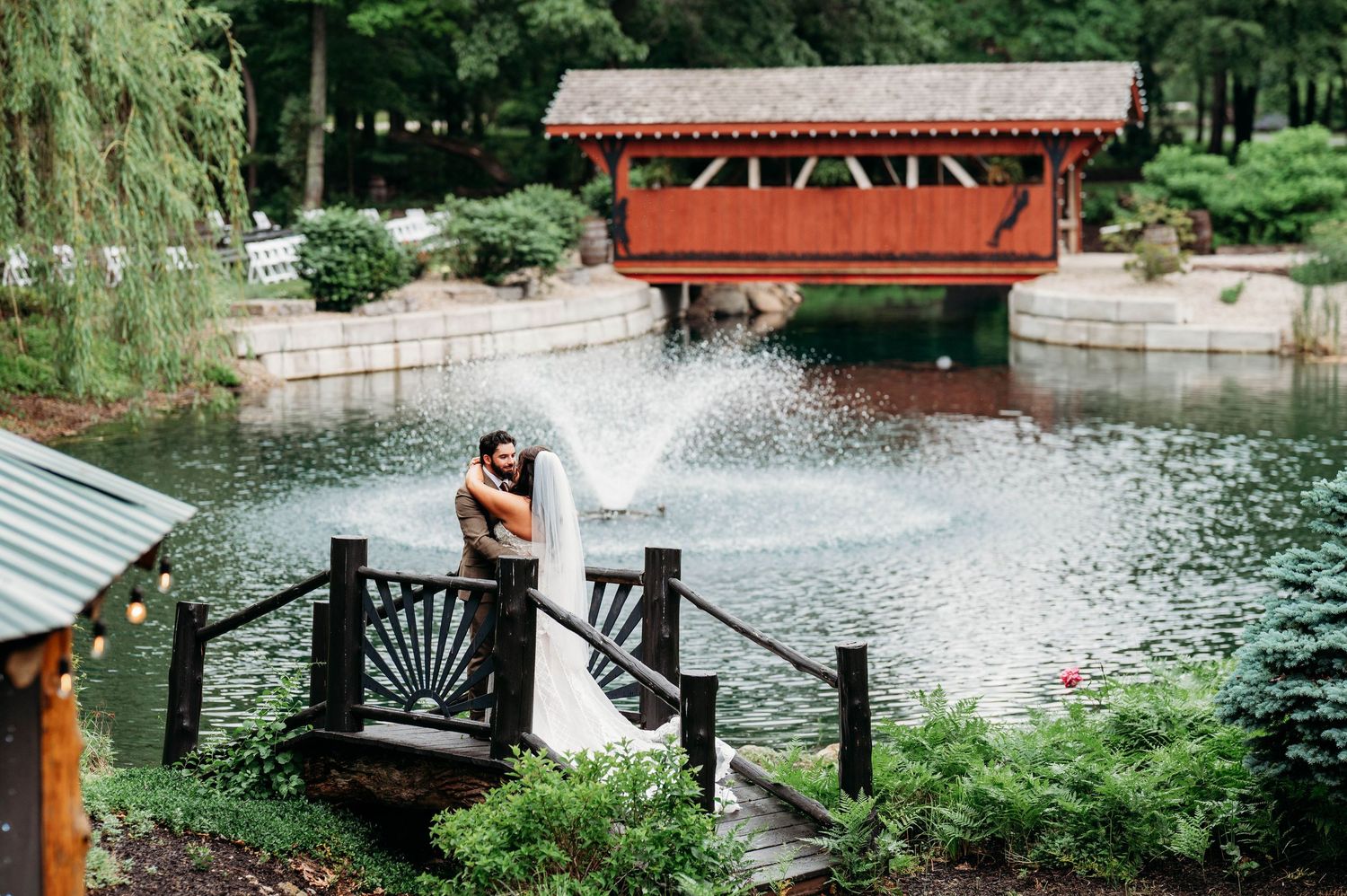 Bride and groom embrace on a wooden bridge over a pond with fountain, red covered bridge in background.