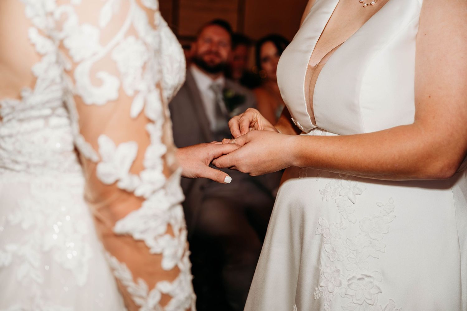 Close-up of hands during wedding ring exchange ceremony showing detailed lace dress and white gown.