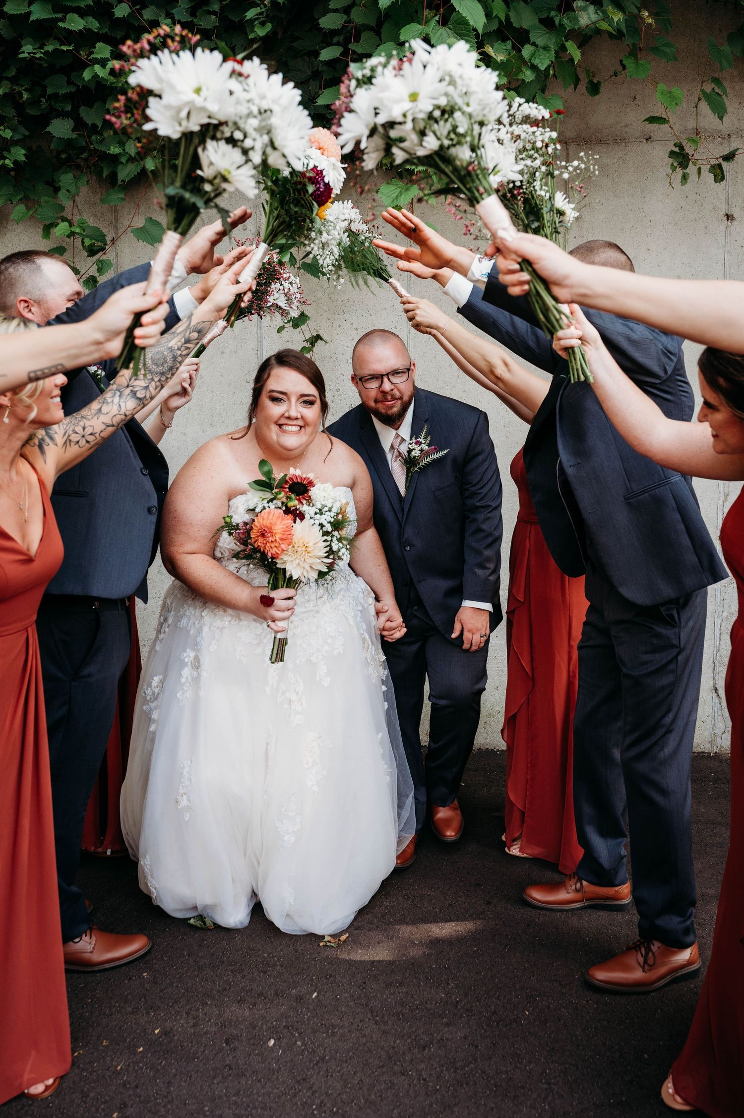 Wedding party in rust-colored dresses and dark suits raise their bouquets in celebration under a floral arch.