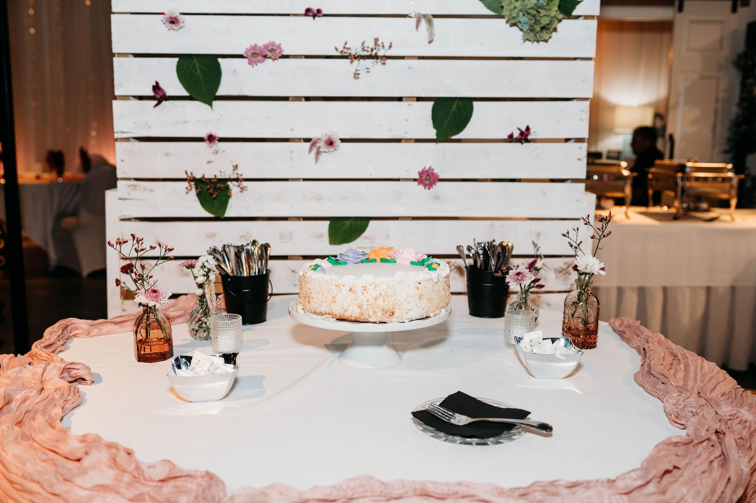Elegant wedding cake table setup with white linens, pink draping, and a wooden slatted backdrop decorated with flowers.