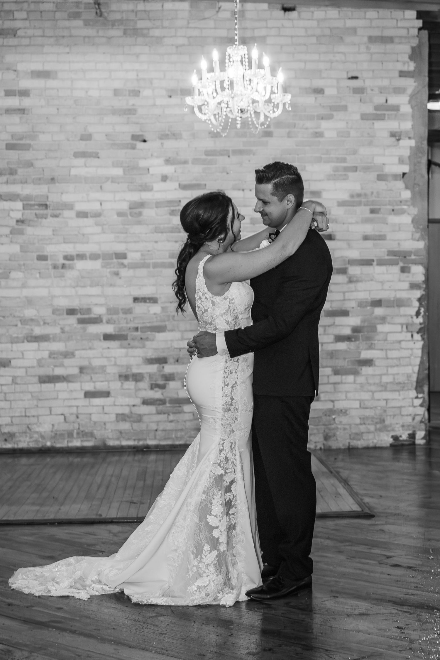 A couple shares their first dance in a rustic brick-walled venue with elegant chandeliers overhead.